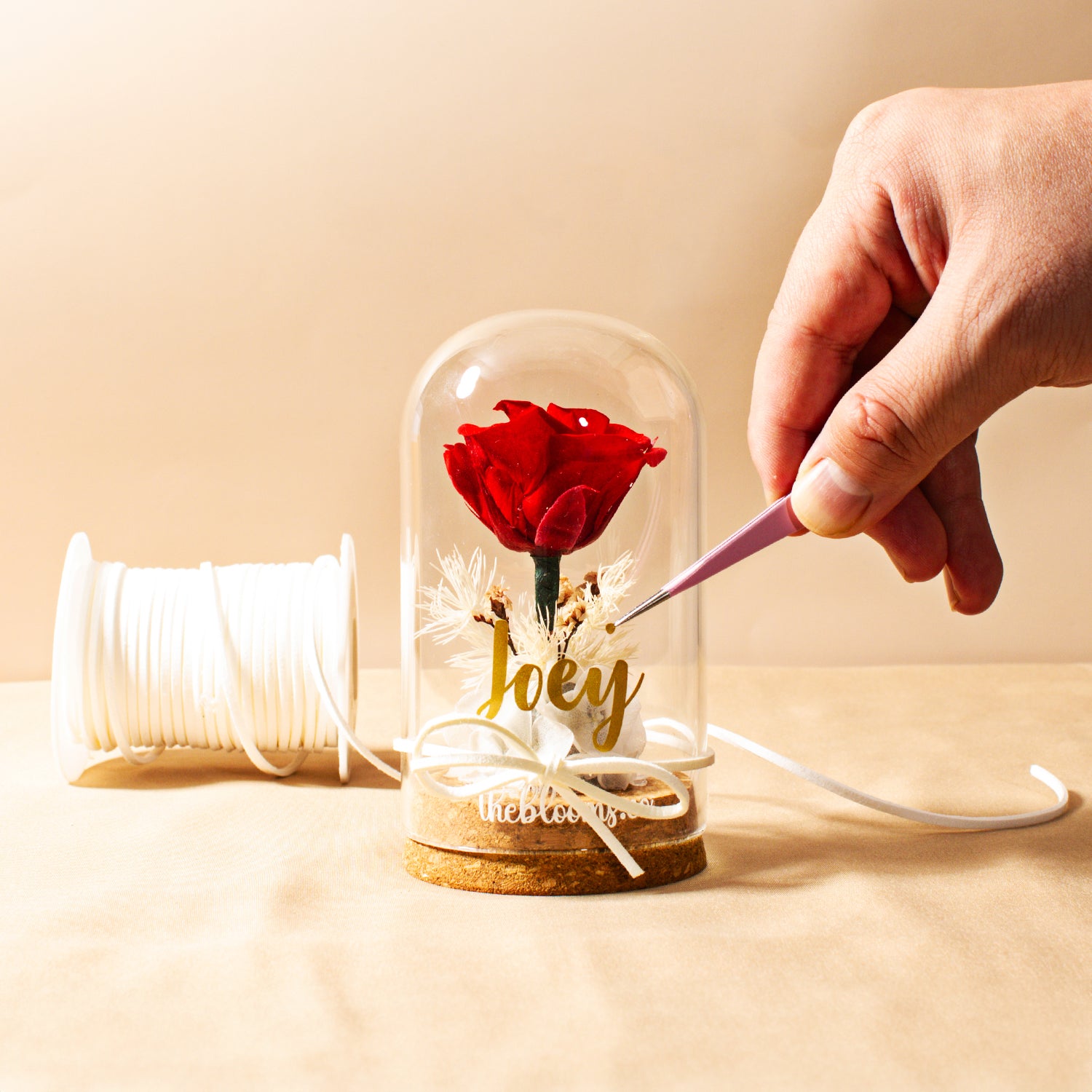 A close-up of a hand with tweezers adjusting a red preserved rose arrangement in a personalized glass dome. A spool of white ribbon is next to the display.