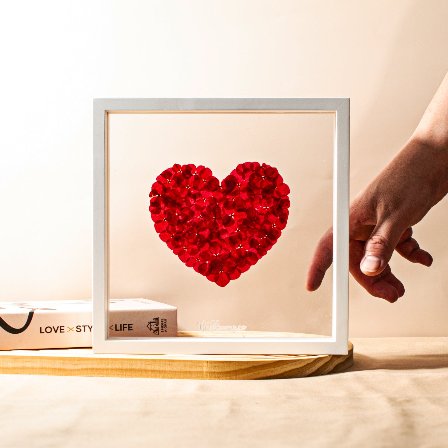 A heart-shaped arrangement of red preserved hydrangea petals in a white frame, with a hand pointing at it on a wooden table.