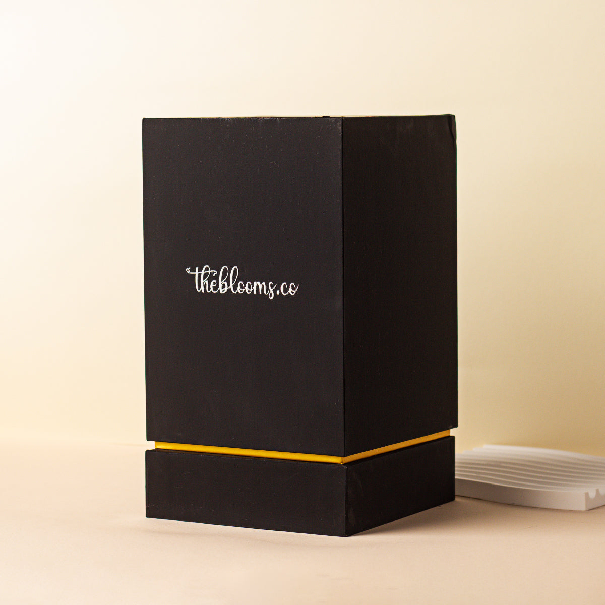 Black and gold branded gift box from Theblooms.co, shown partially open on a beige background.