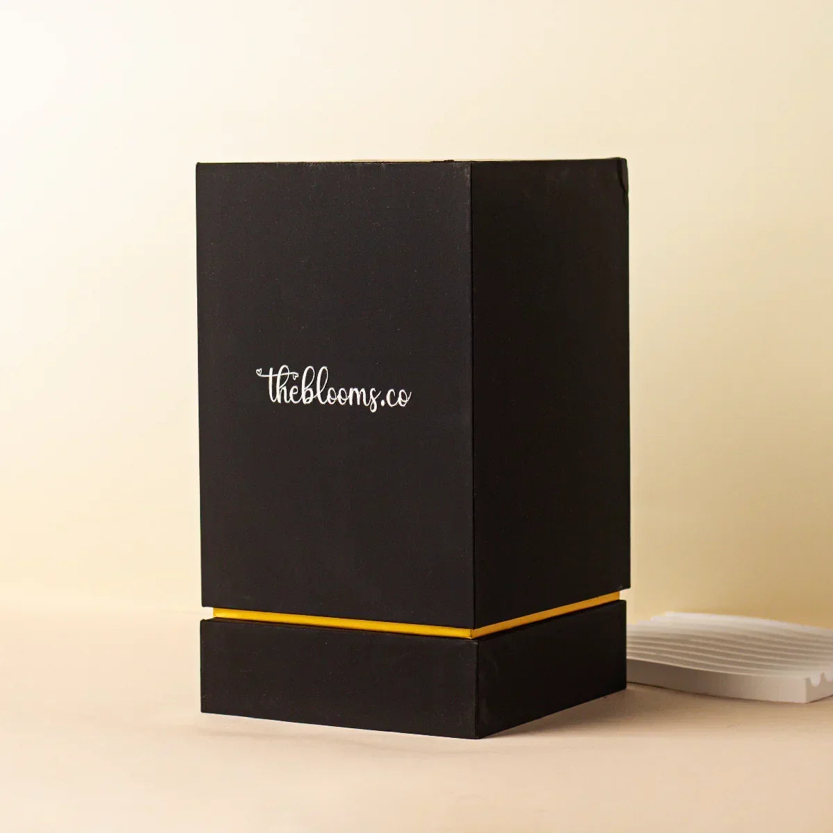 Black and gold branded gift box from Theblooms.co, shown partially open on a beige background.