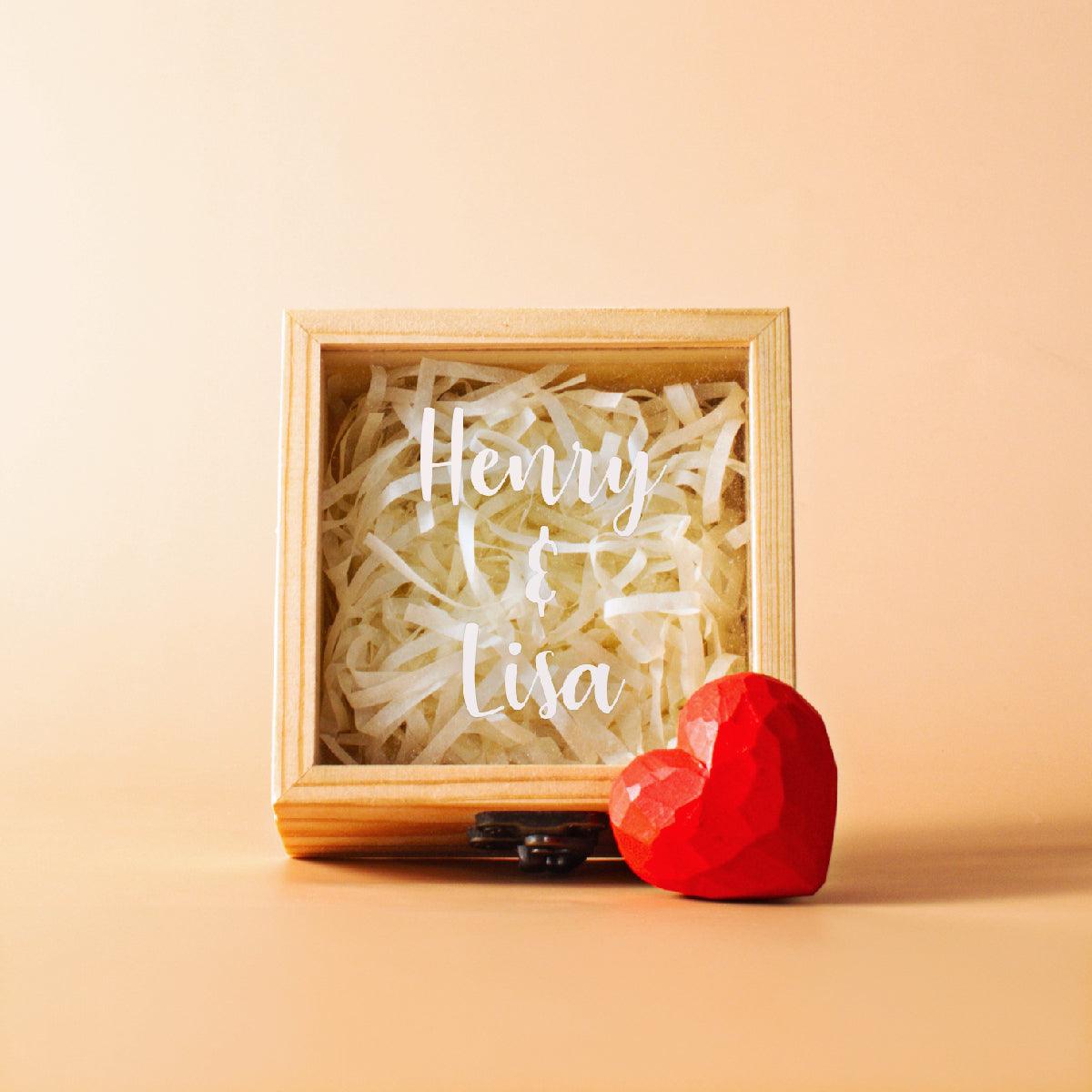 A personalized wooden box with a clear acrylic lid, filled with white shredded paper filler. The lid is customized with the names "Henry & Lisa" in a white script font. A small, faceted red heart sculpture or paperweight rests on the surface next to the box. The background is a plain, light beige color.