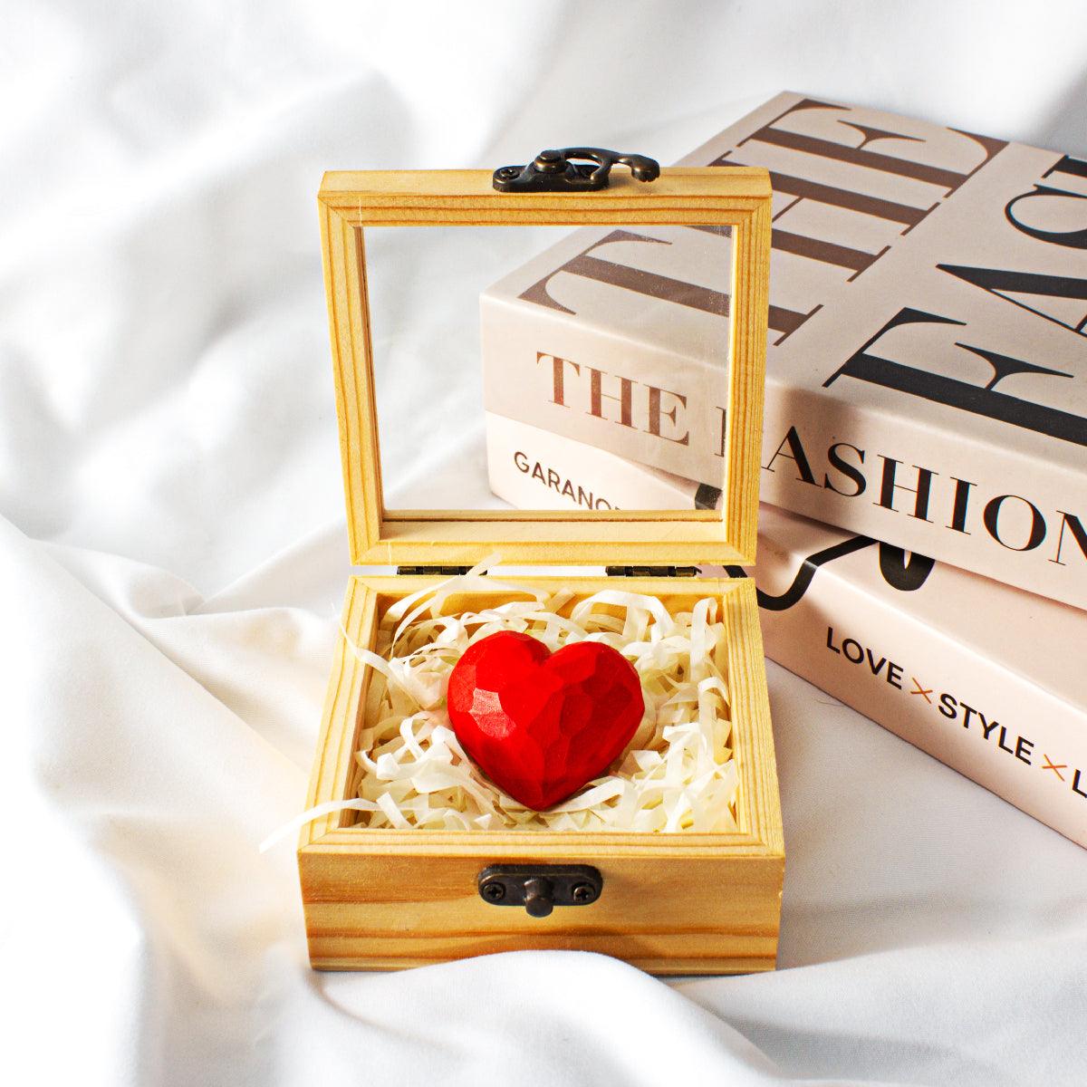 A small, open wooden box with a hinged clear acrylic lid. Inside the box, a red faceted heart sculpture rests on a bed of white shredded paper. The box is set on a white silk or satin fabric background.