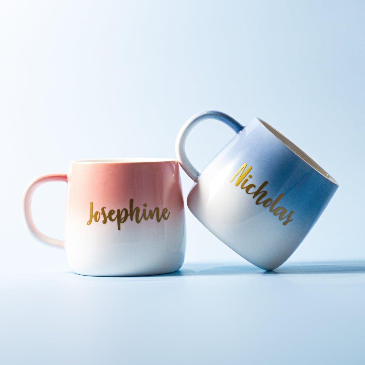 A pair of ceramic coffee mugs with an ombre color design, set against a light blue background. The mug on the left transitions from pink at the top to white at the bottom, and is personalized with the name "Josephine" in gold script. The mug on the right transitions from blue to white, and is personalized with the name "Nicholas" in gold script. The mugs are positioned leaning against each other.