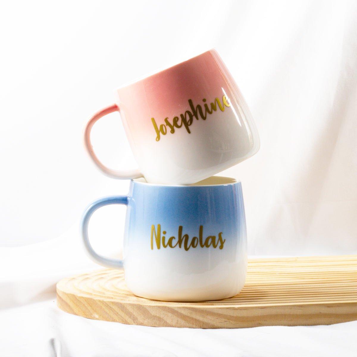 A pair of ceramic ombre mugs, one pink and one blue, stacked on top of each other. The pink mug is on top, personalized with the name "Josephine" in gold script. The blue mug is on the bottom, personalized with the name "Nicholas" in gold script. The mugs are resting on a light wooden board against a soft, white fabric background.