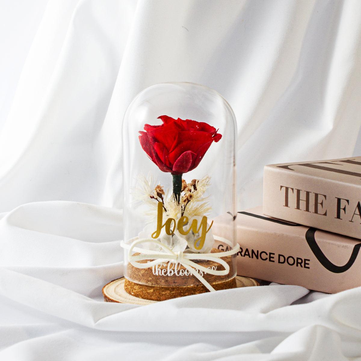 Red preserved rose in glass dome with personalized decal, dried flowers, and ribbon on wooden base. A meaningful romantic gift, styled with books and soft fabric.