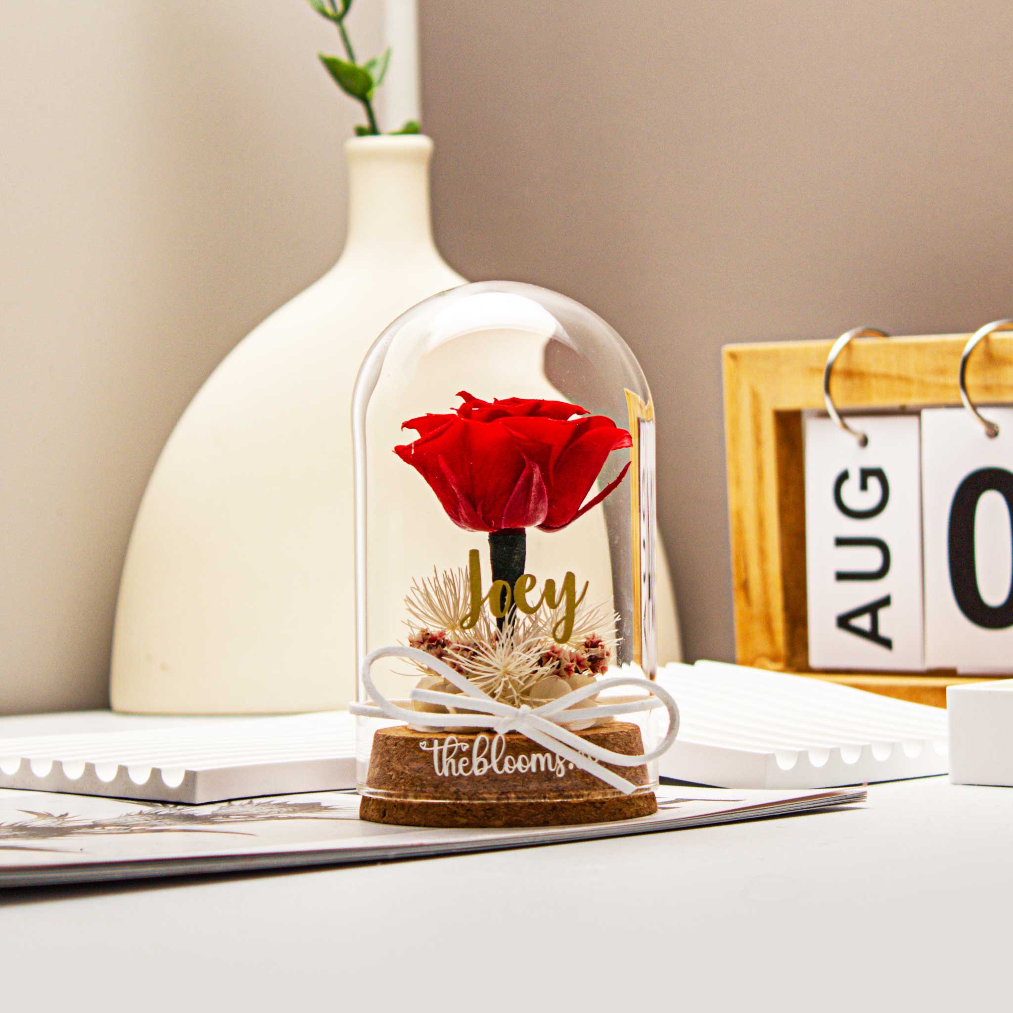 Red preserved rose in glass dome with personalized decal, dried flowers, and ribbon on wooden base. A meaningful romantic gift, styled with books and soft fabric.