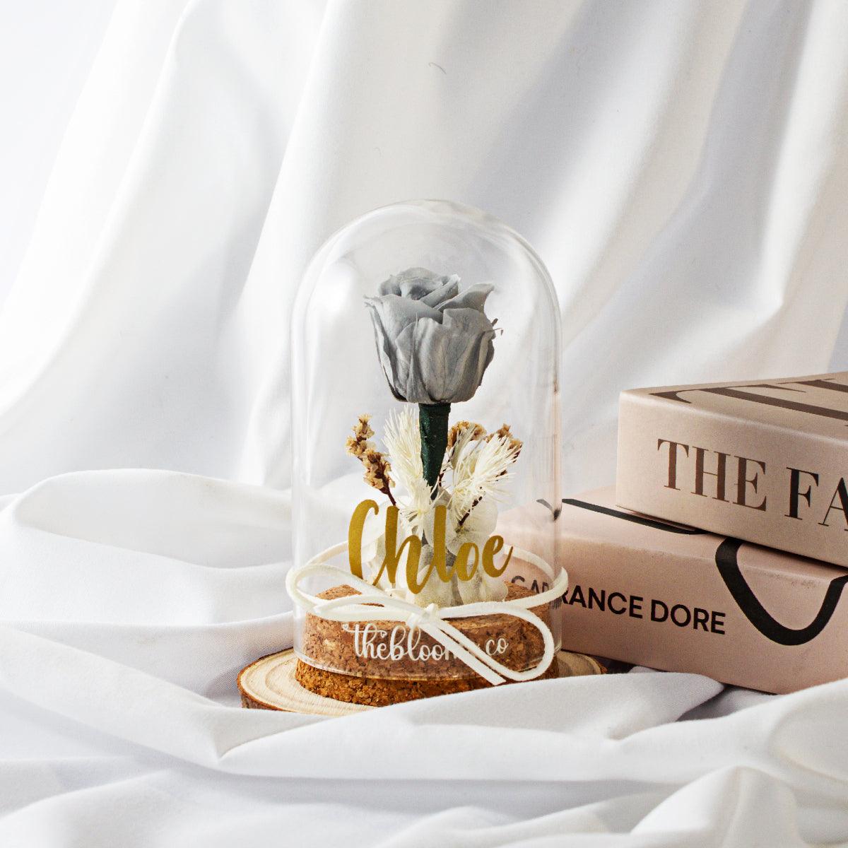 Grey preserved rose in a clear glass dome with white ribbon and dried flower details, displayed on a wooden base. Elegant minimalist floral gift from TheBlooms.co, ideal for modern home decor, anniversaries, or thoughtful occasions.