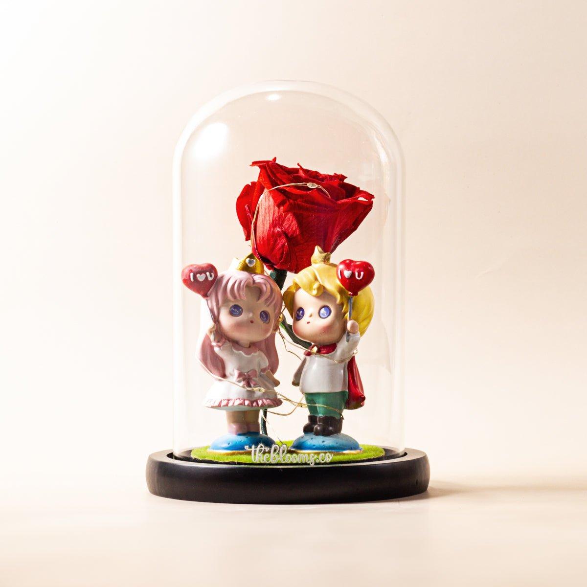 A preserved red rose stands between two small, stylized figurine dolls under a clear glass dome. The figurine on the left has pink hair and holds a heart-shaped balloon, while the one on the right has blonde hair and also holds a heart-shaped balloon. The figurines are standing on a small green base with blue trim. The entire display is on a black wooden base, and the logo "theblooms.co" is visible on the green base. The background is a simple, light beige color.