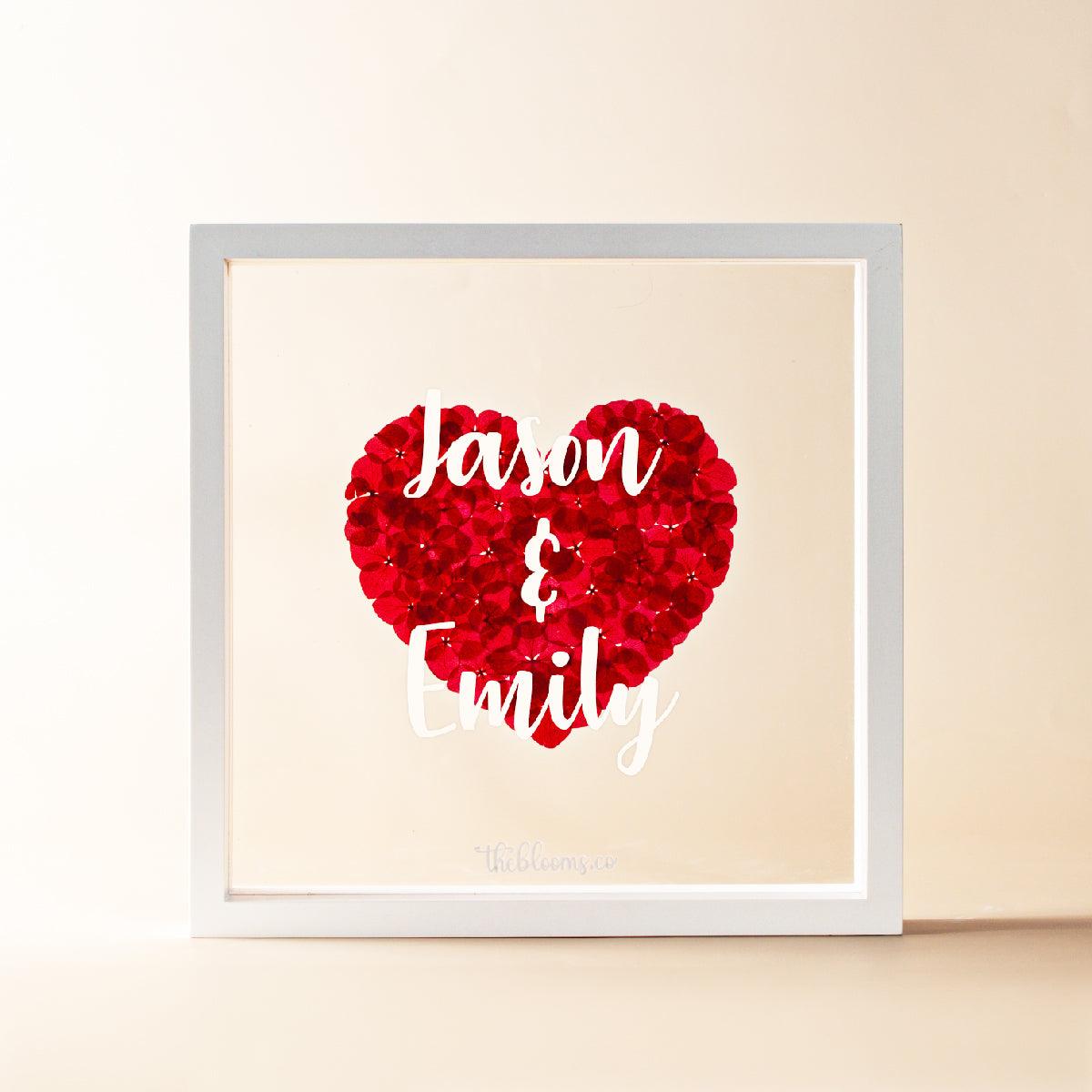 A personalized white wooden shadow box frame containing a heart made of preserved red hydrangea petals. The front glass is customized with the names in white script. The logo "theblooms.co" is at the bottom. The background is a soft, light beige color.