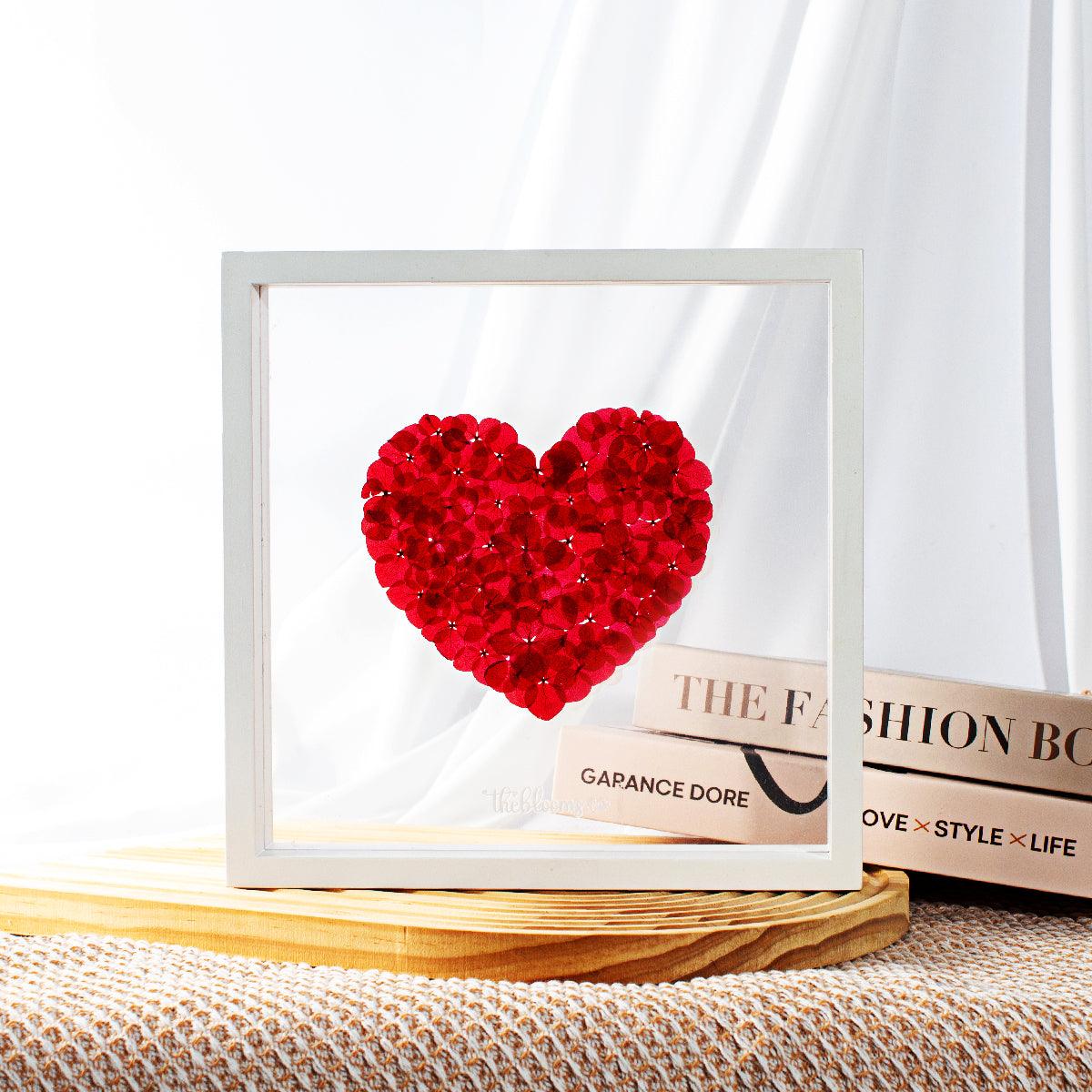 A white wooden shadow box frame containing a heart shape composed of preserved red hydrangea petals. The box is displayed on a light wooden surface with a woven texture underneath.
