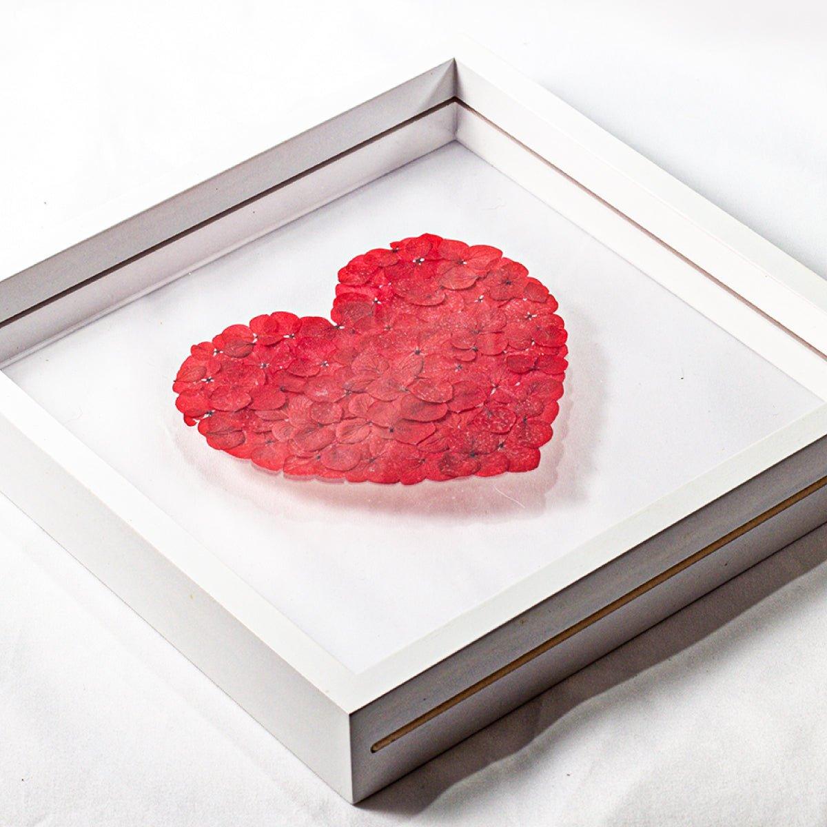 A close-up, high-angle view of a white shadow box frame. Inside, a heart shape is meticulously crafted from small, preserved red hydrangea petals. The object is resting on a white surface, highlighting the detail and three-dimensional quality of the floral arrangement.