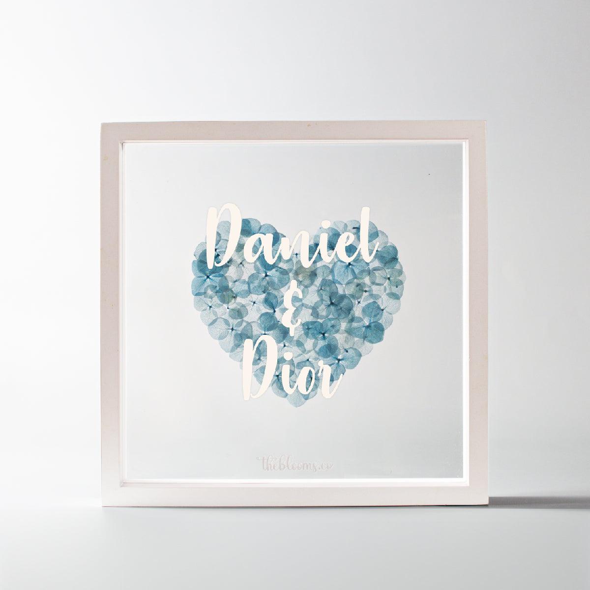 A personalized white wooden shadow box frame containing a heart made of preserved blue hydrangea petals. The front glass is customized with the names "Daniel & Dior" in white script. The small logo "theblooms.co" is at the bottom.