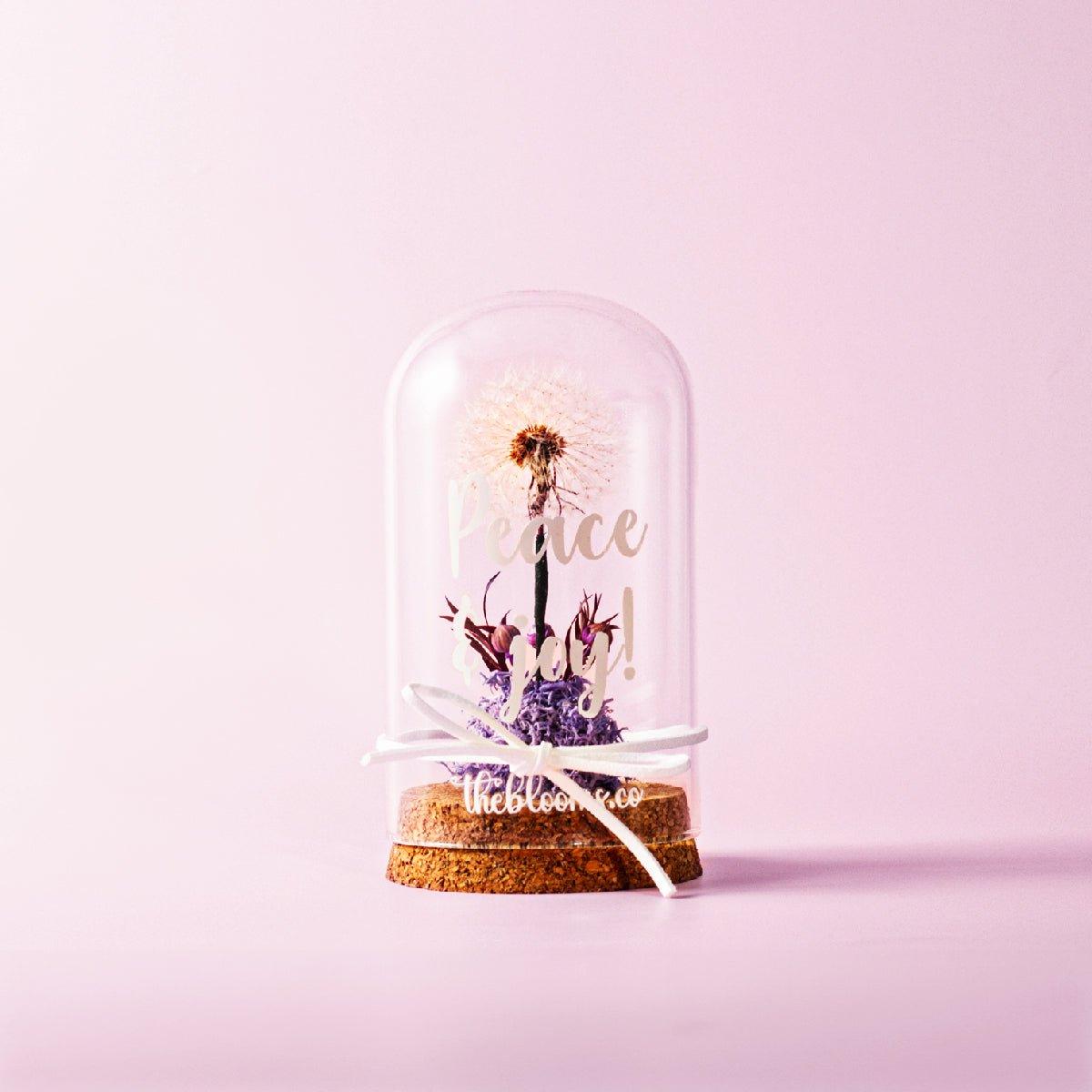 A clear glass dome containing a single dandelion seed head and a small cluster of purple preserved flowers, resting on a cork base. A white ribbon is tied around the base in a bow on the glass dome. The background is a solid light pink color.