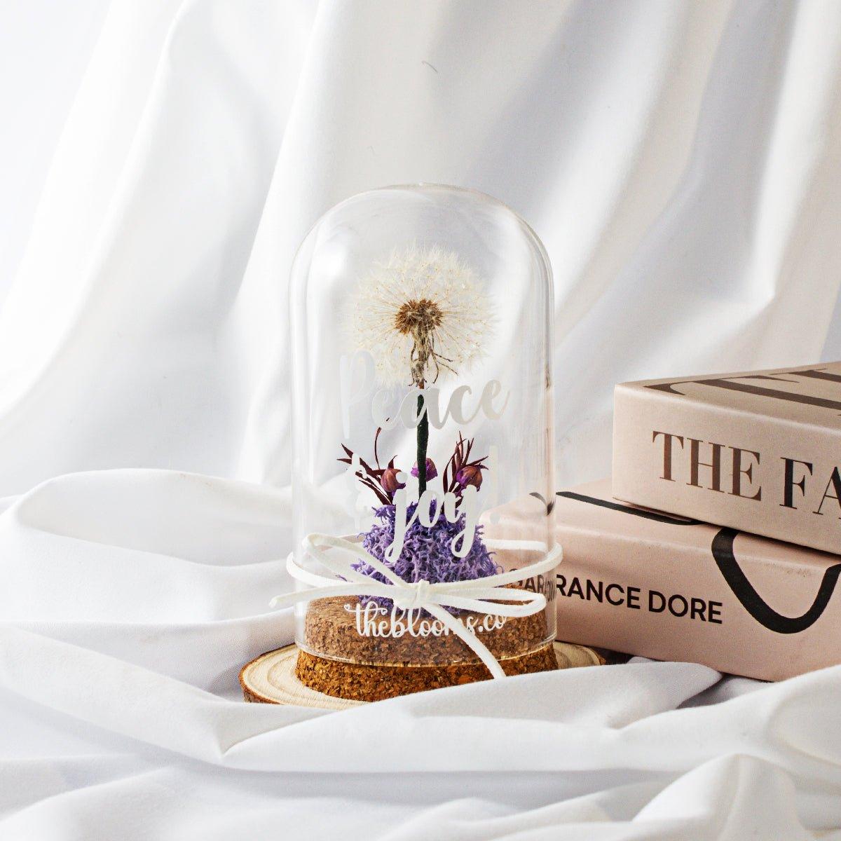 Glass dome with a single, white dandelion puffball and a small cluster of purple preserved flowers inside. The dome is on a round wooden base and tied with a white ribbon.