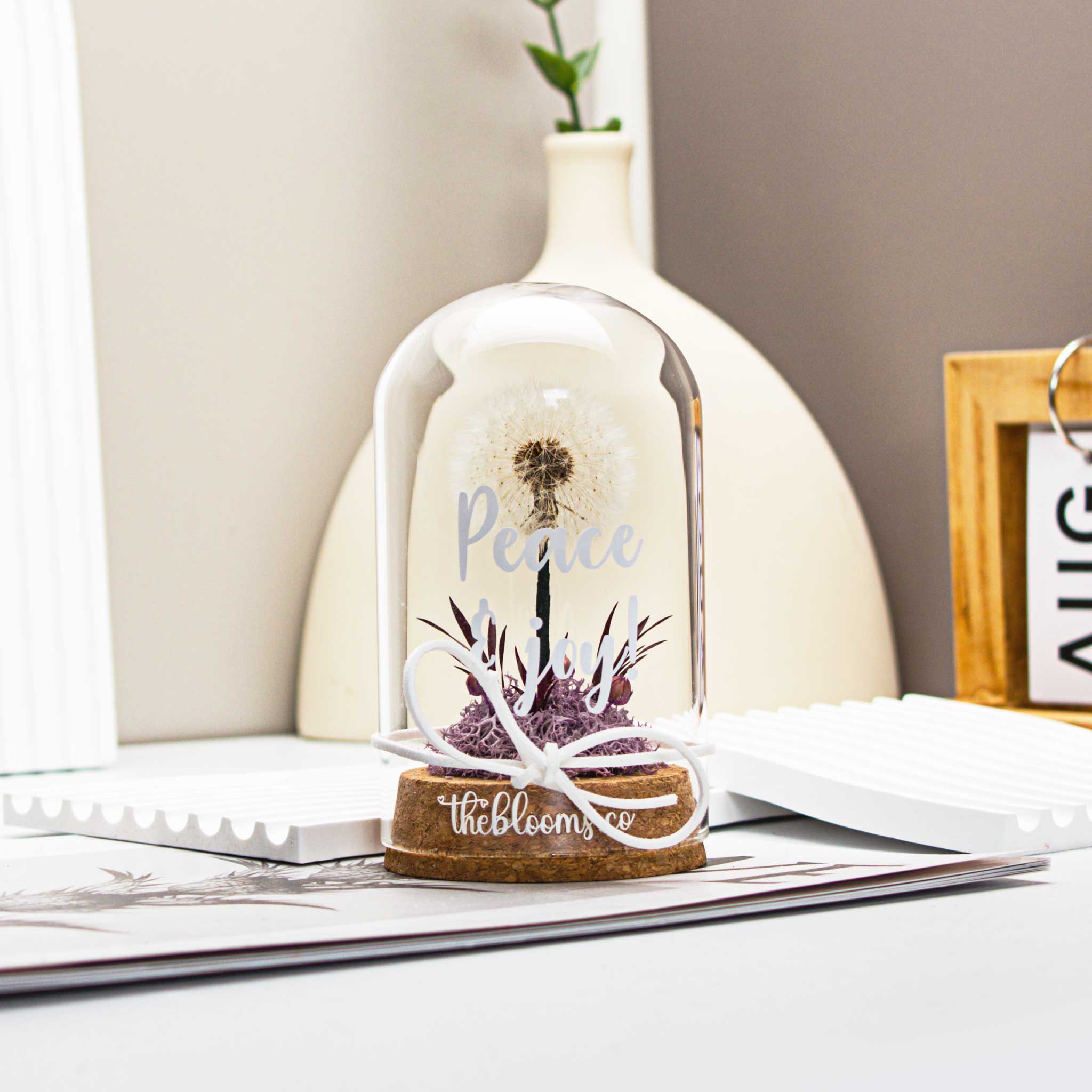 Glass dome with a single, white dandelion puffball and a small cluster of purple preserved flowers inside. The dome is on a round wooden base and tied with a white ribbon.