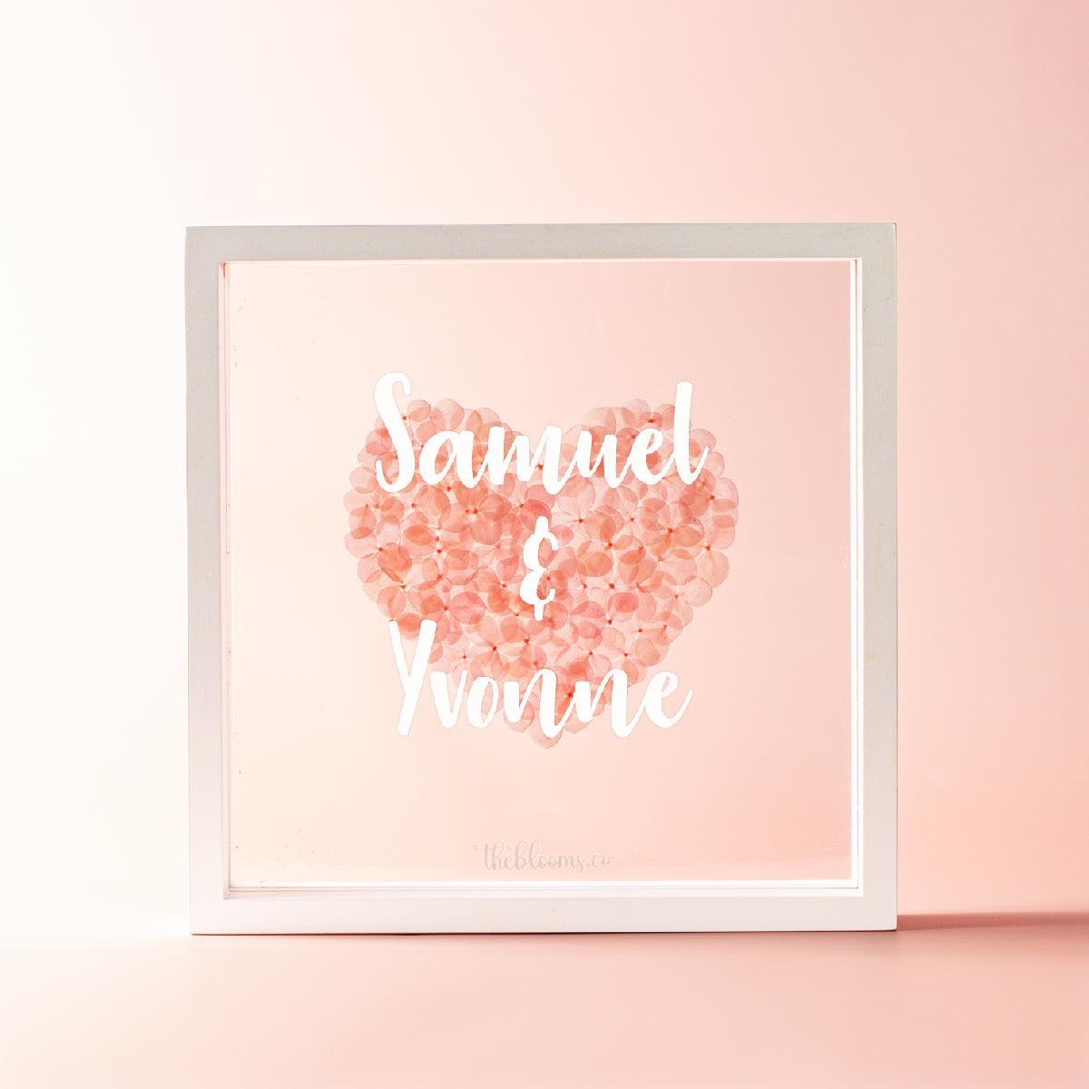A personalized white wooden shadow box frame containing a heart made of preserved pink hydrangea petals. The front glass is customized with the names in white script. The logo "theblooms.co" is at the bottom. The background is a soft, light pink color.