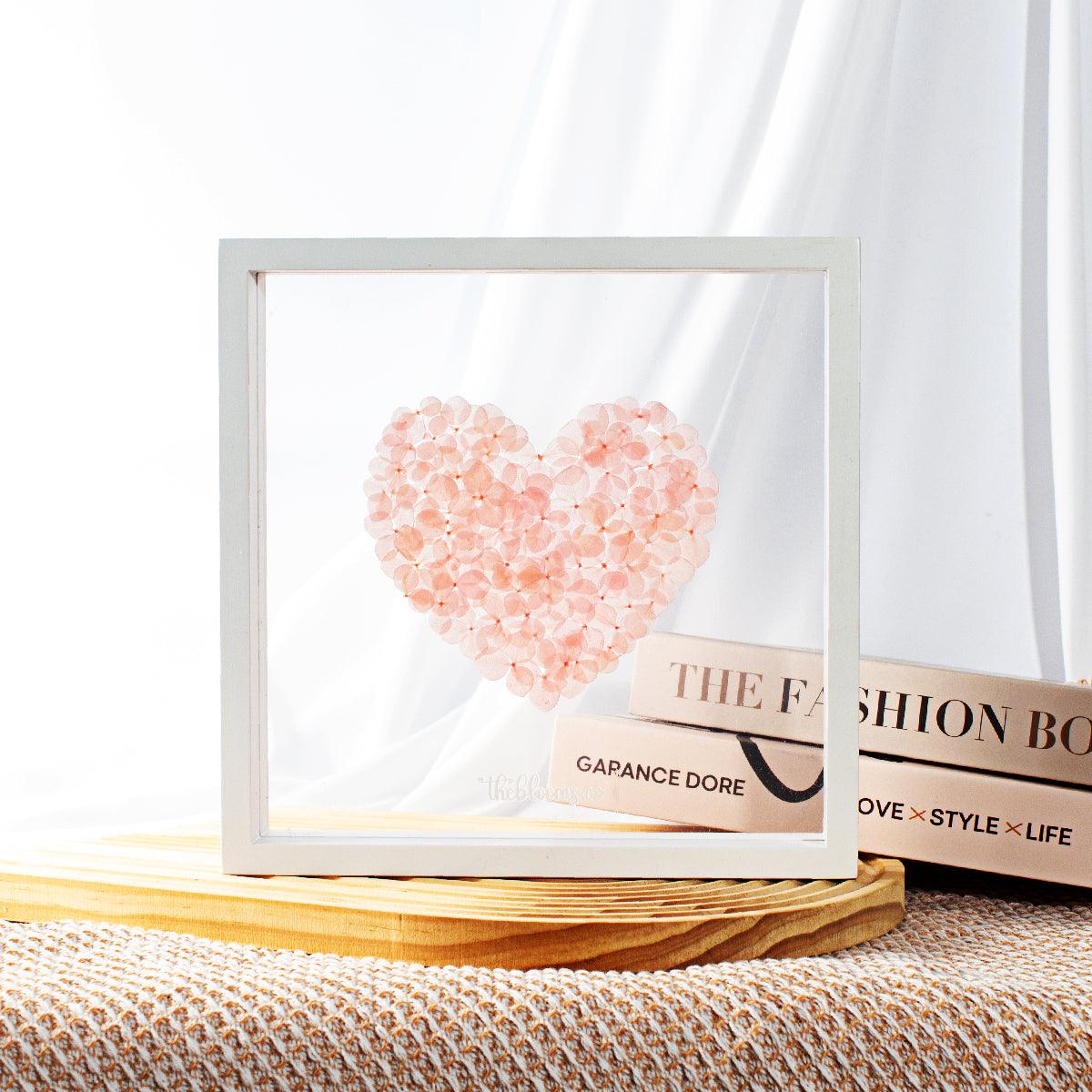 A white wooden shadow box frame containing a heart shape composed of preserved pink hydrangea petals. The box is displayed on a light wooden surface with a woven texture underneath.