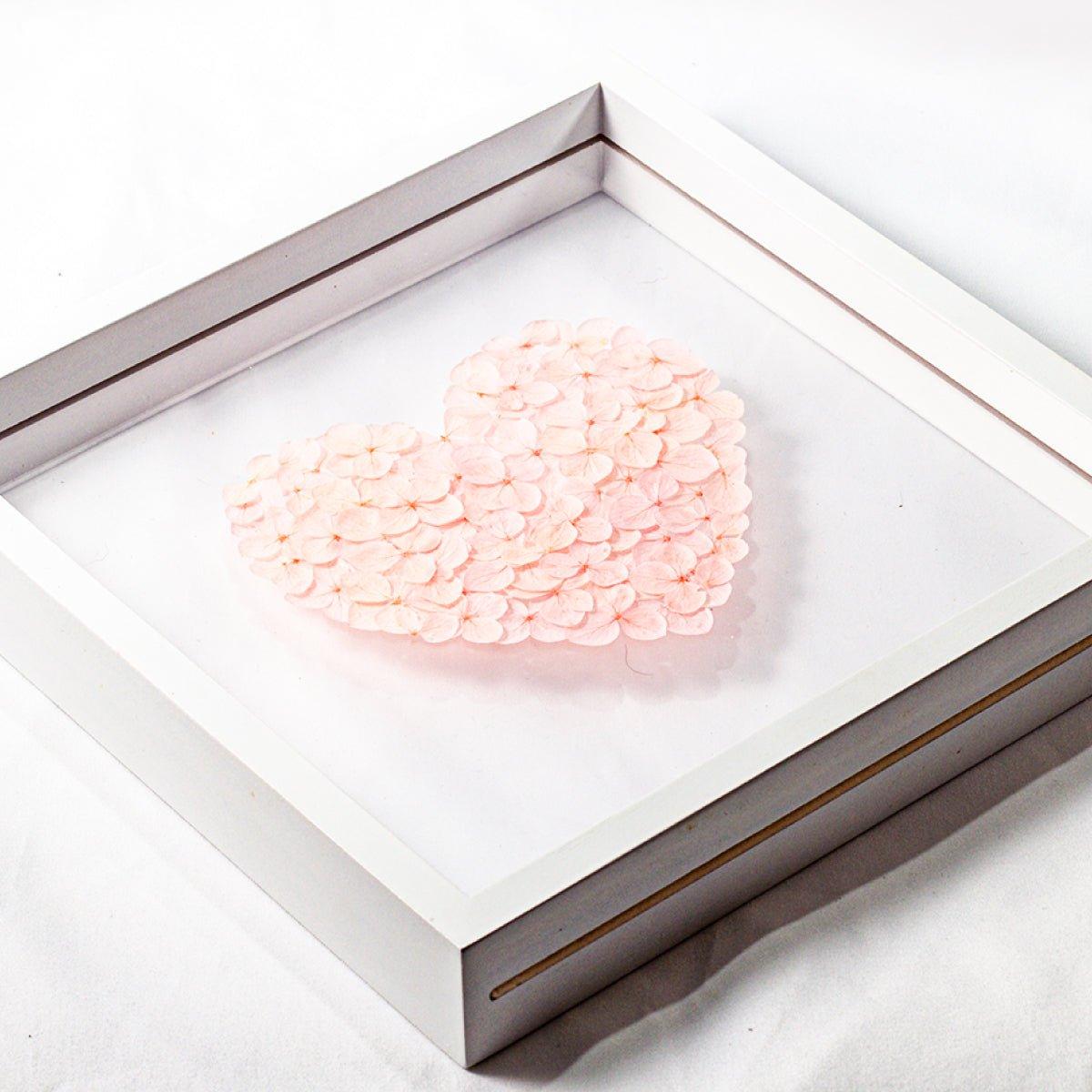 A close-up, high-angle view of a white shadow box frame. Inside, a heart shape is meticulously crafted from small, preserved pink hydrangea petals. The object is resting on a white surface, highlighting the detail and three-dimensional quality of the floral arrangement.