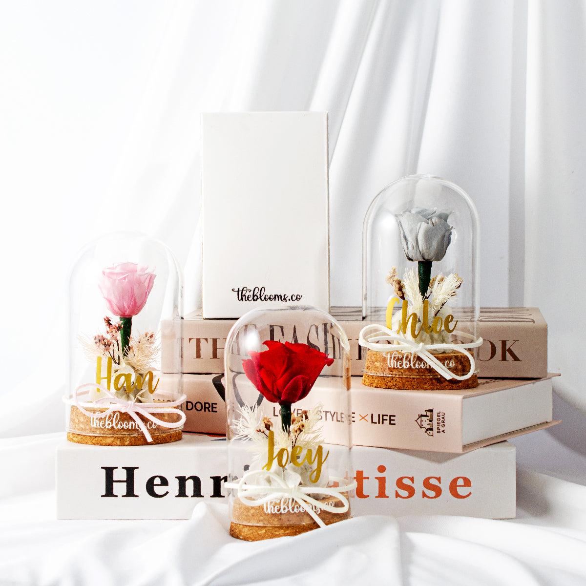Preserved roses in elegant glass domes with name personalization, dried floral accents, and satin ribbons. Displayed on stacked books, ideal as romantic gifts from TheBlooms.co.