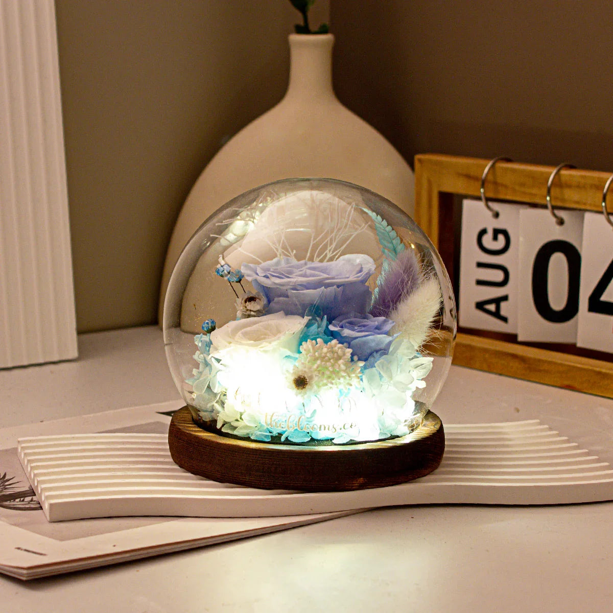 Preserved blue and white roses in a glass dome with LED lights on a wooden base, styled on a desk with a calendar.