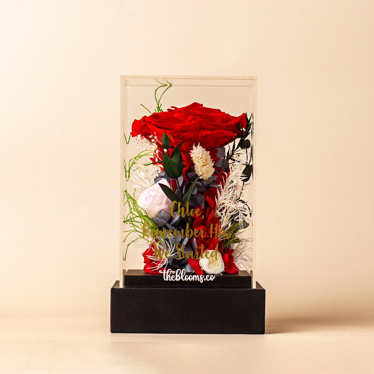 Front view of a personalized preserved floral arrangement in a clear acrylic box, featuring a large red rose and various white and grey flowers. The box has a custom gold inscription and sits on a black base.