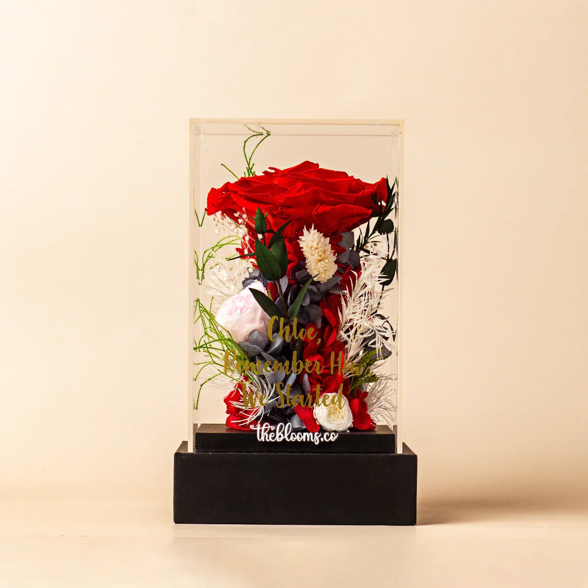 Front view of a personalized preserved floral arrangement in a clear acrylic box, featuring a large red rose and various white and grey flowers. The box has a custom gold inscription and sits on a black base.