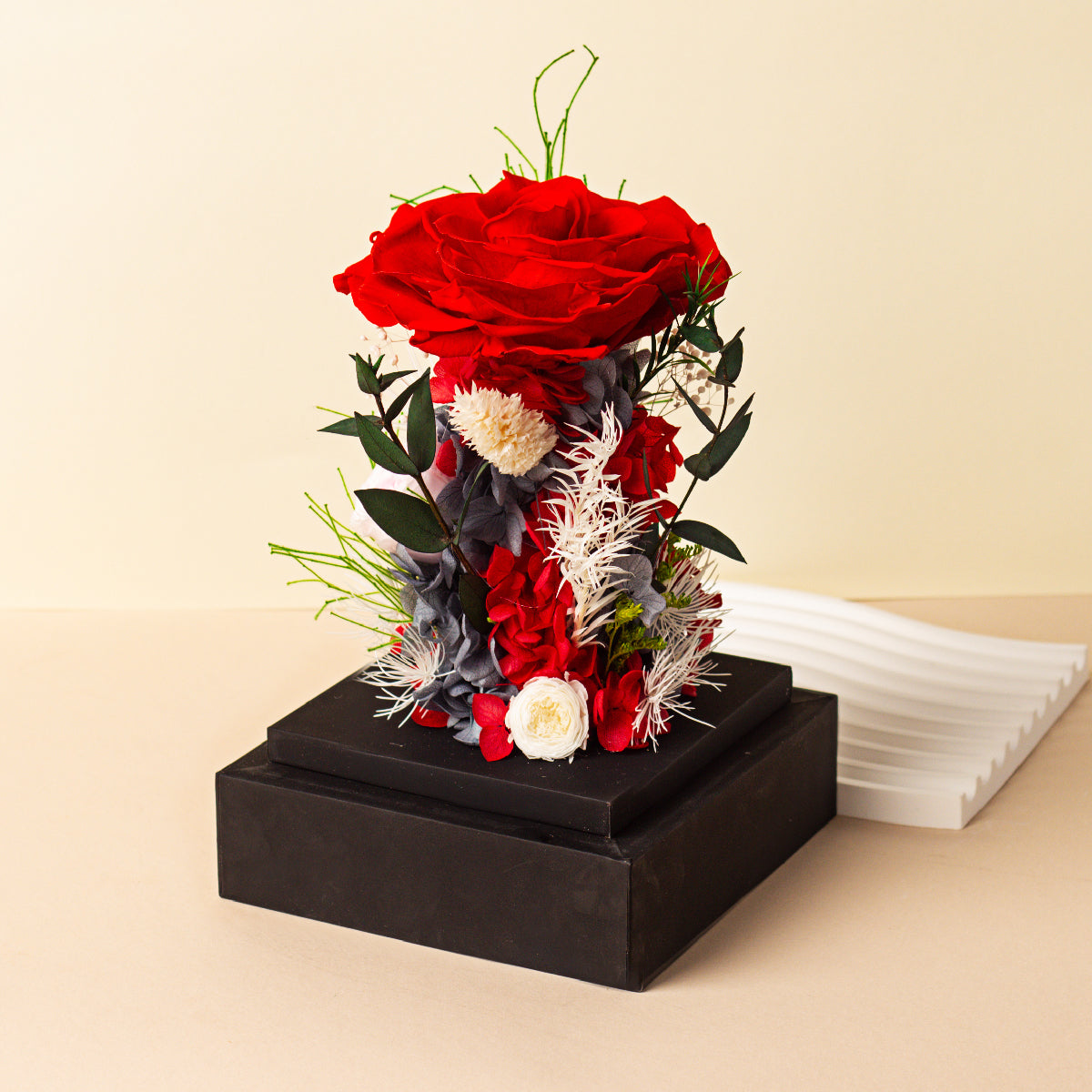 A red preserved rose floral arrangement with white and other accent flowers on a acrylic box, with a white wavy decorative object to the side.