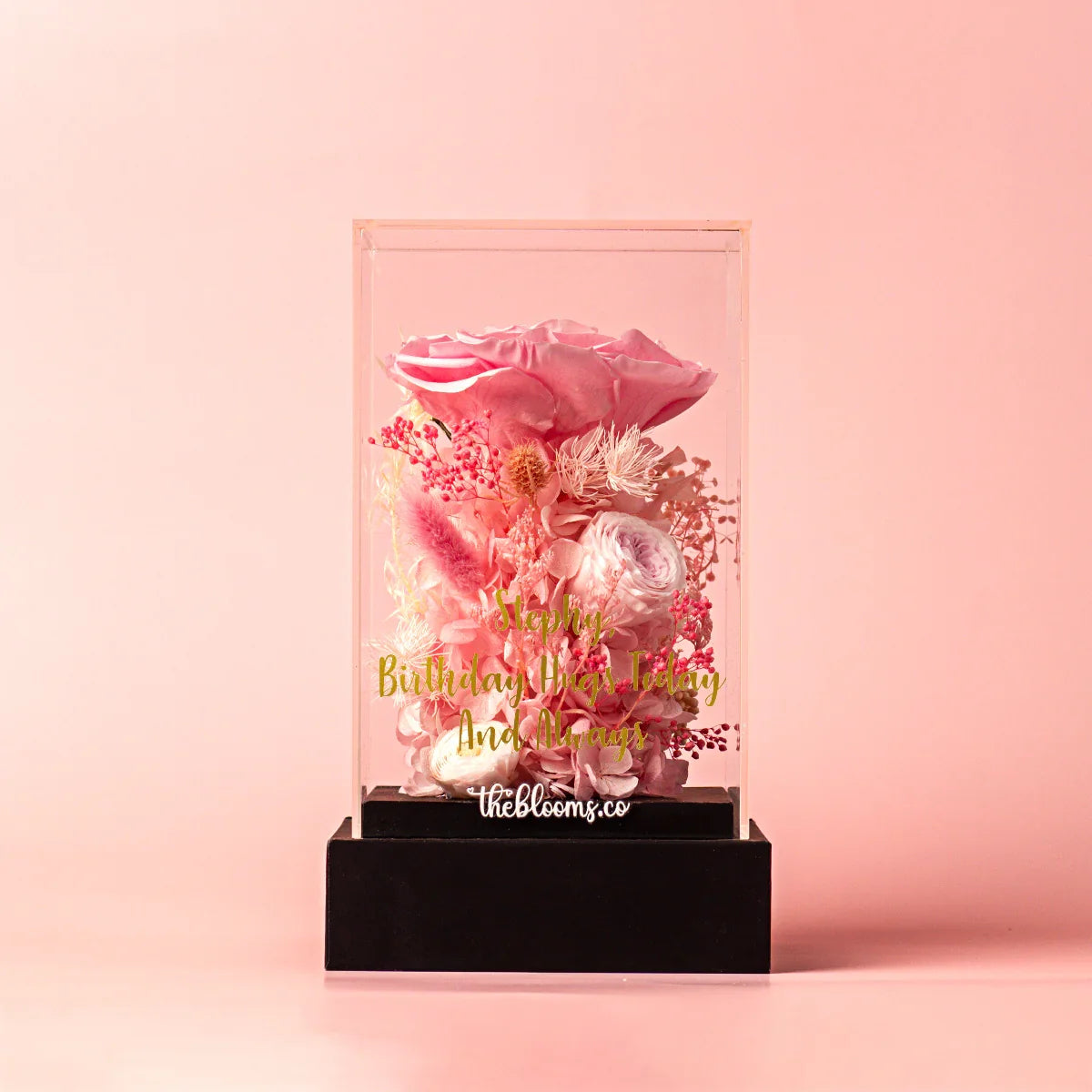 Front view of a personalized preserved floral arrangement in a clear acrylic box, featuring a large pink rose and various pink dried flowers. The box has a custom gold inscription and sits on a black base.