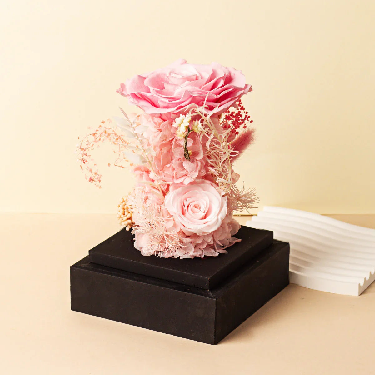 A preserved floral arrangement featuring a large pink rose and various pink dried flowers, displayed on a tiered black wooden base next to a white decorative object.