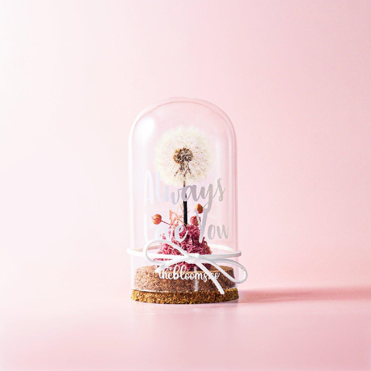 A clear glass dome with a single dandelion puffball and a small cluster of pink preserved flowers and berries inside. The dome is on a cork base with a white ribbon tied around on the glass dome. The background is a solid light pink color.