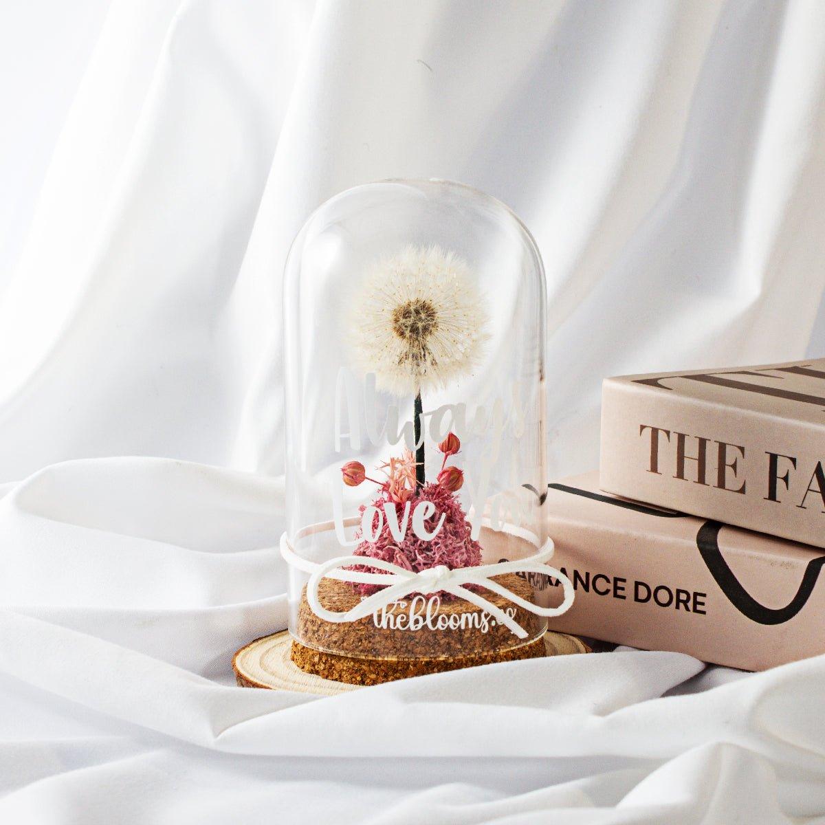 Glass dome with a single, white dandelion puffball and a small cluster of Pink preserved flowers inside. The dome is on a round wooden base and tied with a white ribbon.