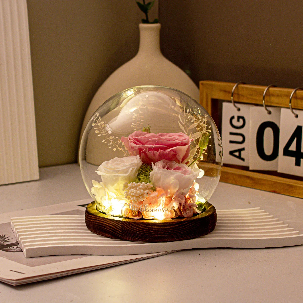 The Enchanted Rose preserved pink and white flowers in a lighted glass dome on a wooden base, styled with a desk calendar.