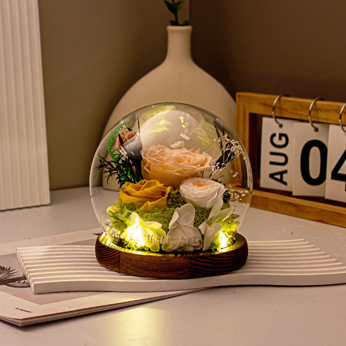 The Secret Garden preserved yellow and champagne roses in a lighted glass globe with green moss, styled on a desk for home decor.