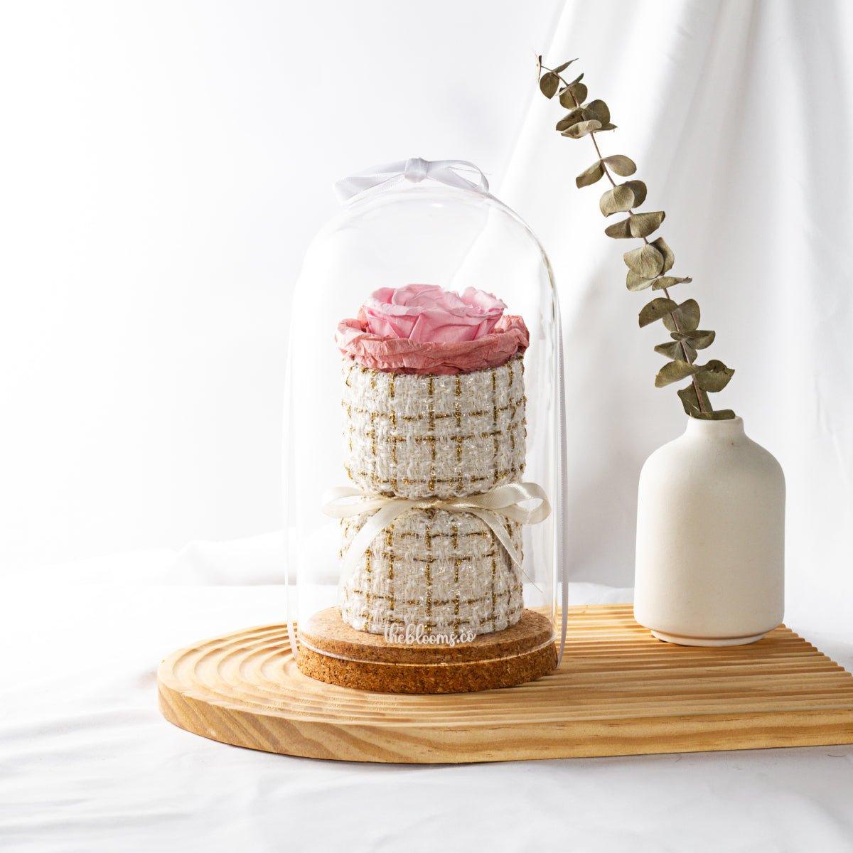 A tall glass dome encases a light pink preserved rose on a two-tiered, white and gold tweed-like base. A white ribbon is tied at the top and another around the middle of the base. The arrangement sits on a round cork base, which is on a light-colored wooden cutting board. Next to it, in a small white ceramic vase, are dried eucalyptus leaves. The background is a soft white fabric.