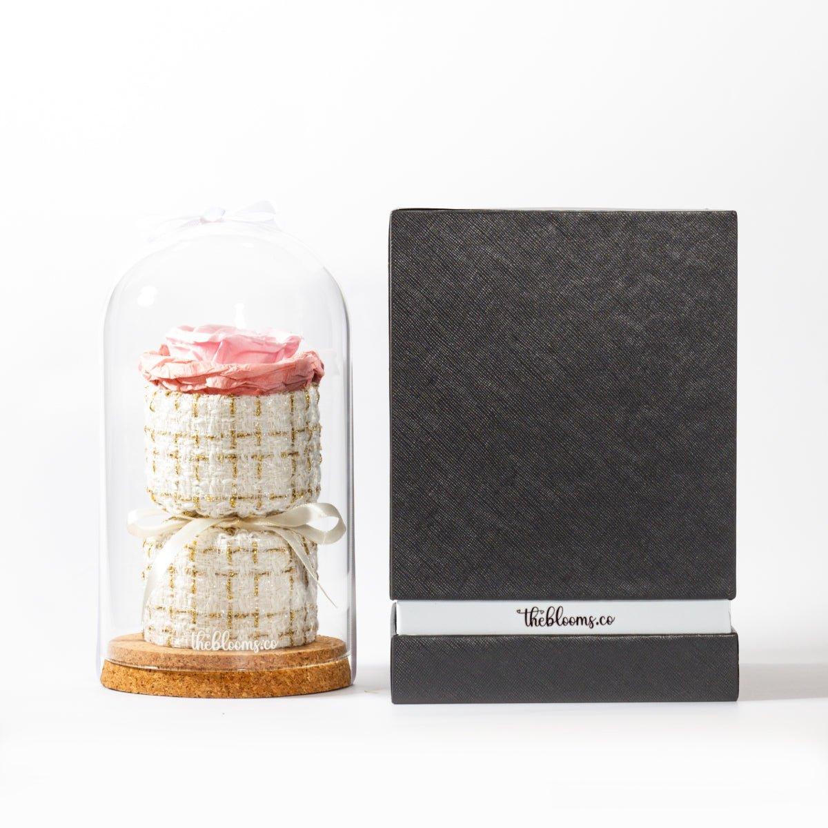 A pink preserved rose in a glass dome on a cork base, with a two-tiered, white and gold tweed base inside. Next to it stands a tall, black textured gift box with a white band around the bottom that reads "theblooms.co". The two items are shown side-by-side on a white surface with a simple white background.