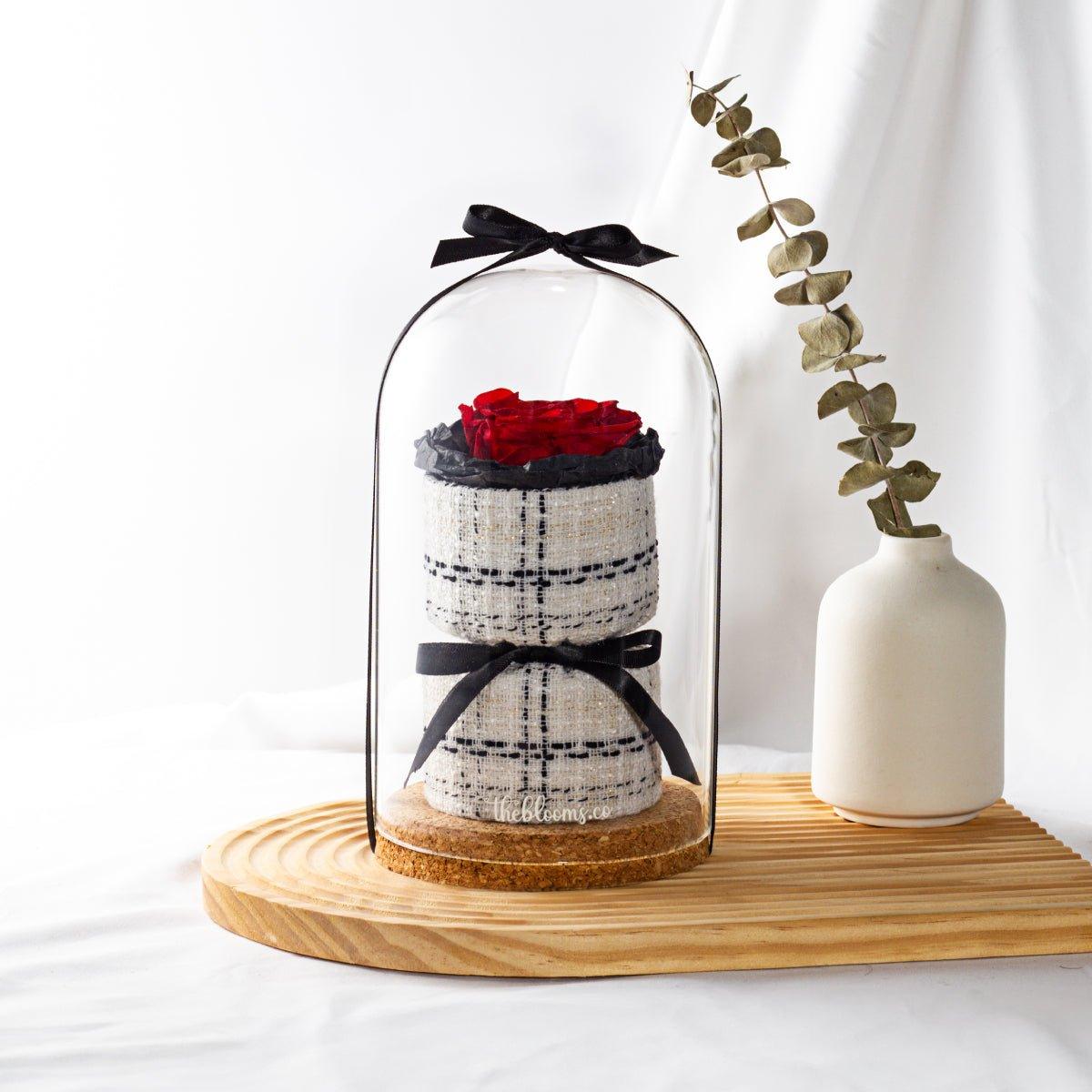 A tall glass dome encases a red preserved rose on a two-tiered, white and black plaid tweed-like base. A black ribbon is tied at the top and another around the middle of the base. The arrangement sits on a round cork base, which is on a light-colored wooden cutting board. Next to it, in a small white ceramic vase, are dried eucalyptus leaves. The background is a soft white fabric.