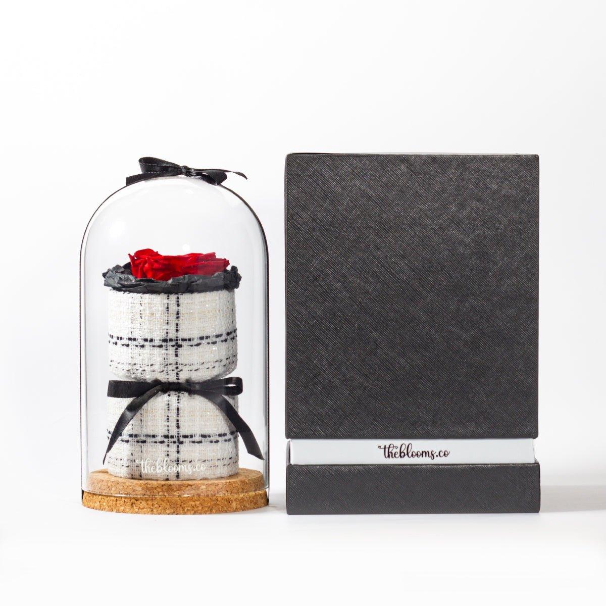 A red preserved rose in a glass dome on a cork base, with a two-tiered, white and black plaid tweed base inside. Next to it stands a tall, black textured gift box with a white band around the bottom that reads "theblooms.co". The two items are shown side-by-side on a white surface with a simple white background.