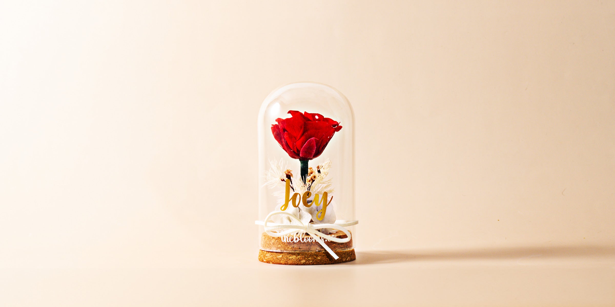 A single, vibrant red rose is preserved under a clear glass dome, sitting on a round wooden base. The rose is surrounded by what appear to be small white filler flowers. The name "Joey" is written in a elegant gold script on the glass, and a small white ribbon is tied around the base of the dome. The background is a plain, light beige color.