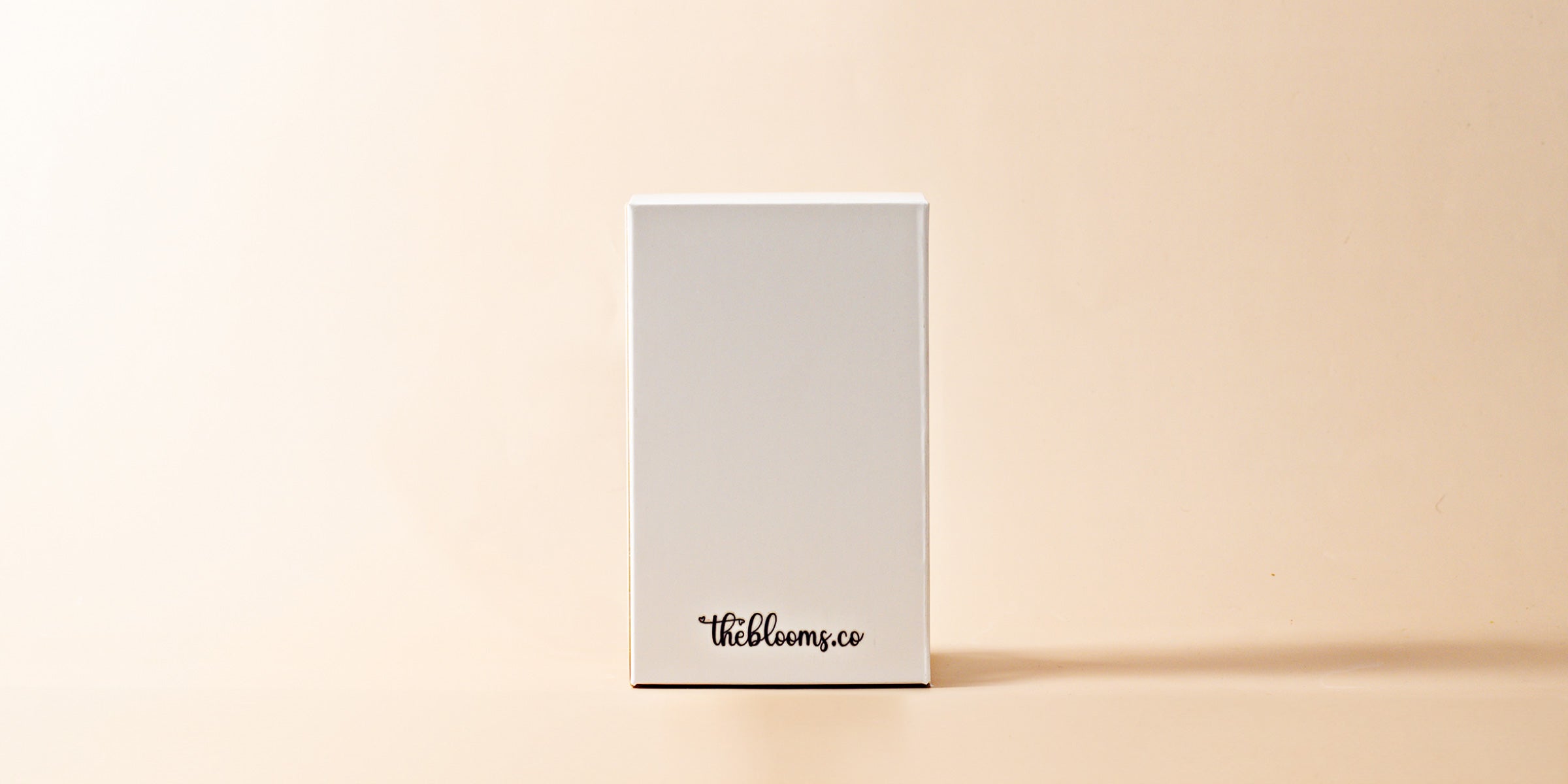 A rectangular, off-white gift box stands upright against a plain, light beige background. The bottom front of the box has a small logo or text in black, which reads "theblooms.co". The box appears simple and clean, with no other prominent features.