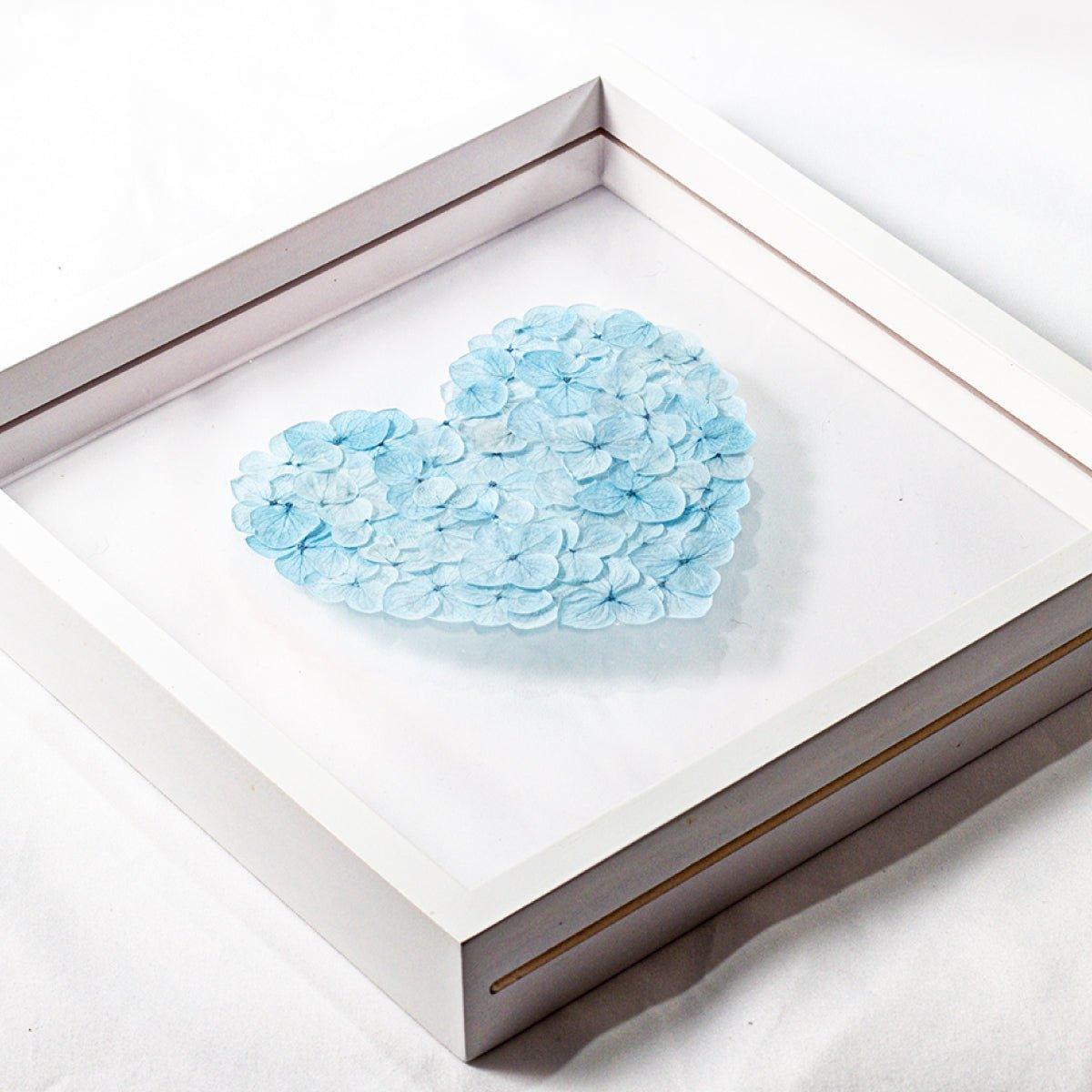 A close-up, high-angle view of a white shadow box frame. Inside, a heart shape is meticulously crafted from small, preserved blue hydrangea petals. The object is resting on a white surface, highlighting the detail and three-dimensional quality of the floral arrangement.