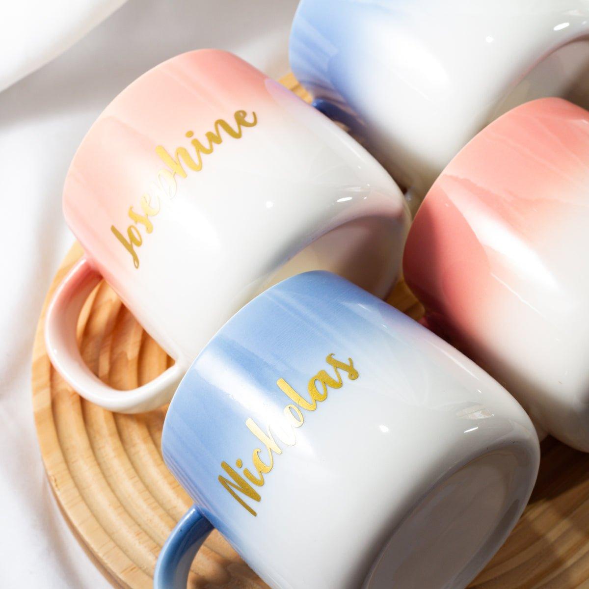 A close-up, high-angle view of two pairs of personalized ceramic ombre mugs. One pair is pink and white, with the name "Josephine" in gold script. The other pair is blue and white, with the name "Nicholas" in gold script. The mugs are artfully arranged on a round, light-colored wooden board against a soft, white fabric background.