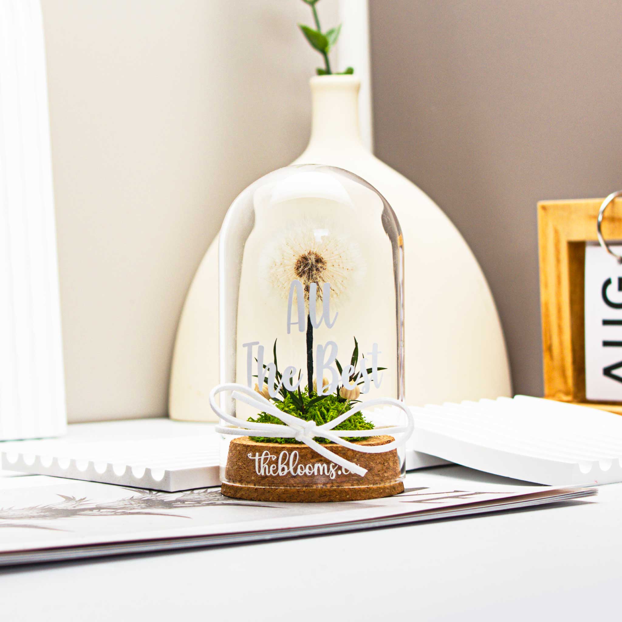 Glass dome with a single, white dandelion puffball and a small cluster of  green preserved flowers inside. The dome is on a round wooden base and tied with a white ribbon.