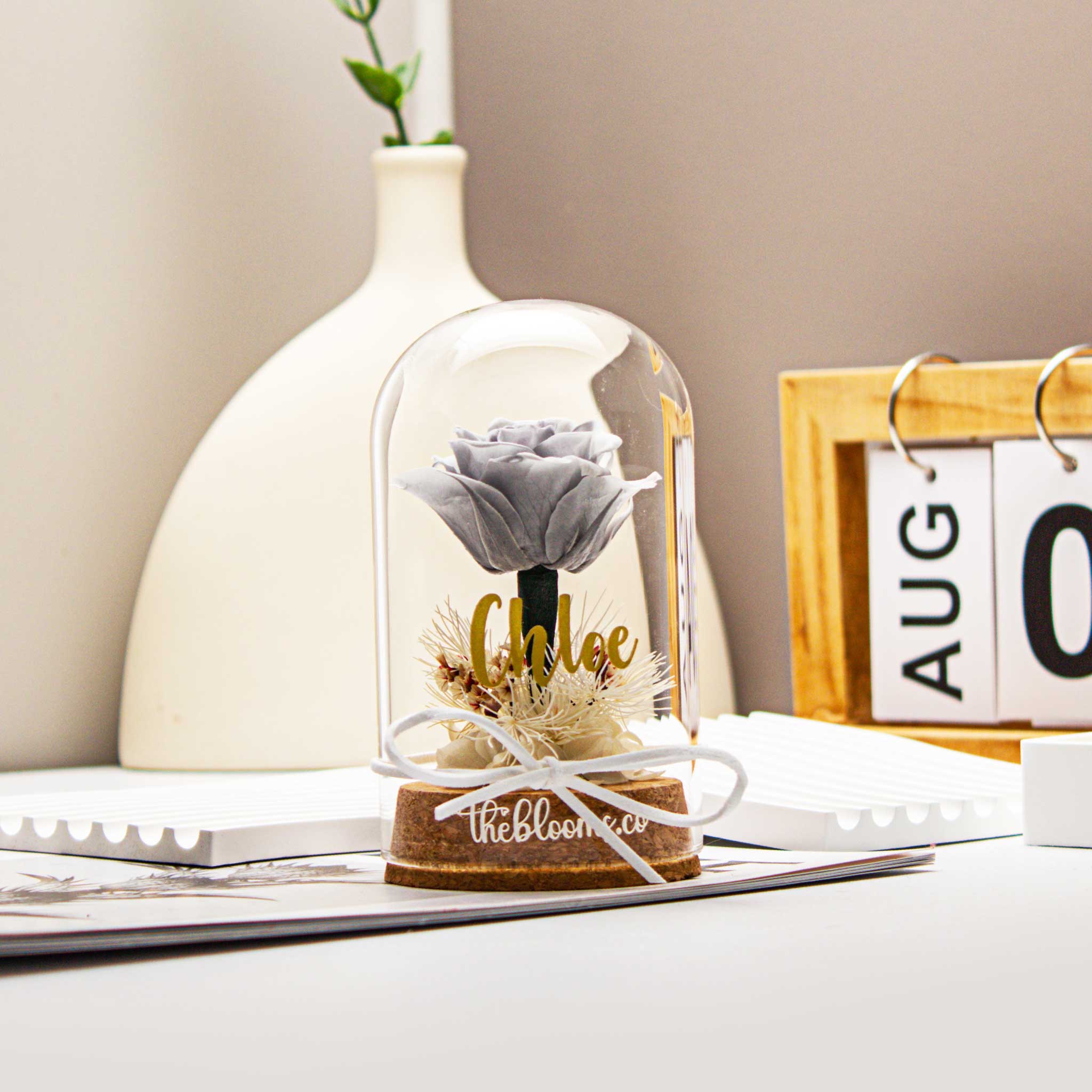 Grey preserved rose in a clear glass dome with white ribbon and dried flower details, displayed on a wooden base. Elegant minimalist floral gift from TheBlooms.co, ideal for modern home decor, anniversaries, or thoughtful occasions.