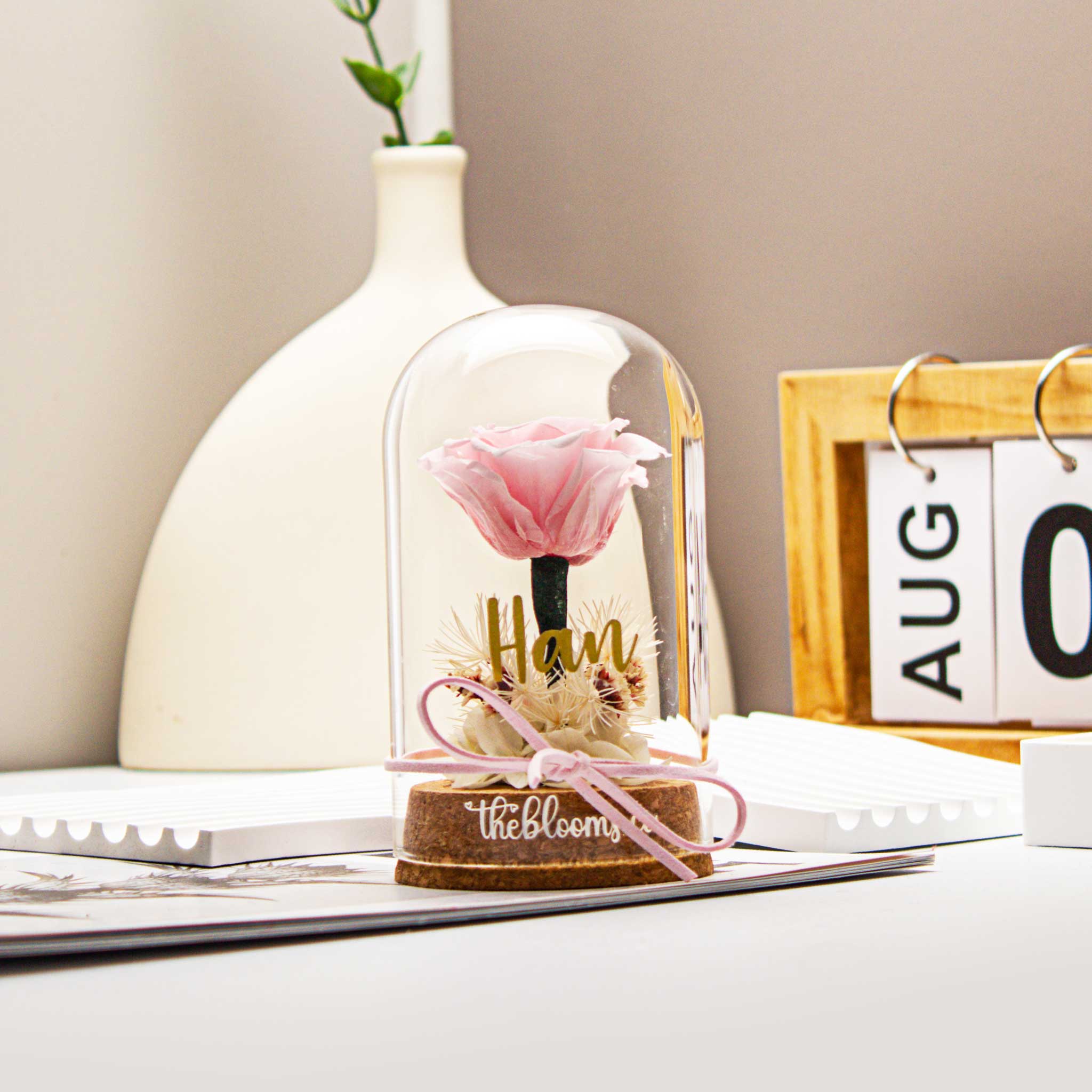 Pink preserved rose in a clear glass dome with ribbon and dried flowers, set against a soft pink background. Elegant personalized floral gift from TheBlooms.co, ideal for birthdays, anniversaries, or romantic occasions.