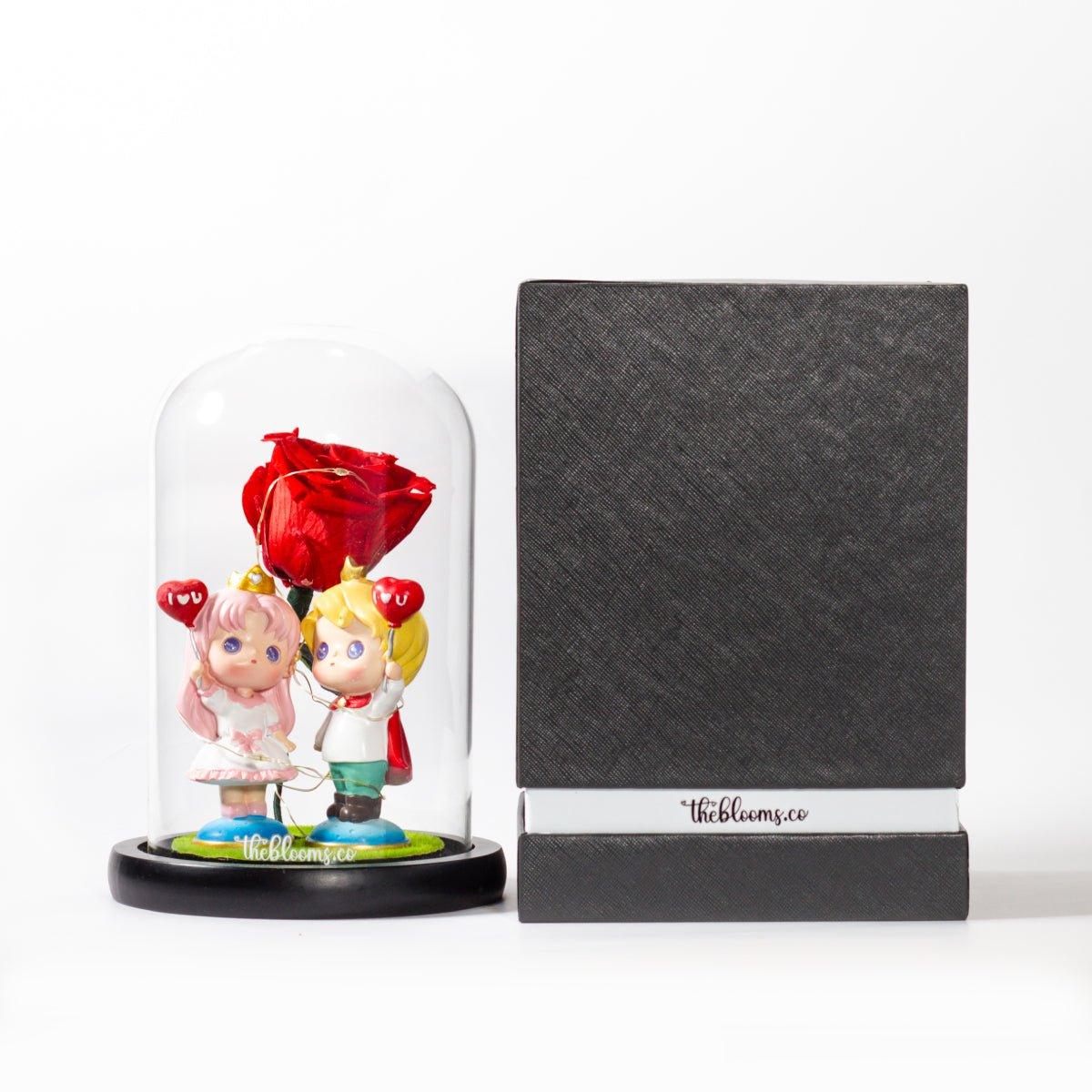 A preserved red rose and two figurines in a glass dome on a black wooden base. Next to it stands a tall, black textured gift box with a white band around the bottom that reads "theblooms.co". The two items are shown side-by-side on a white surface with a simple white background.