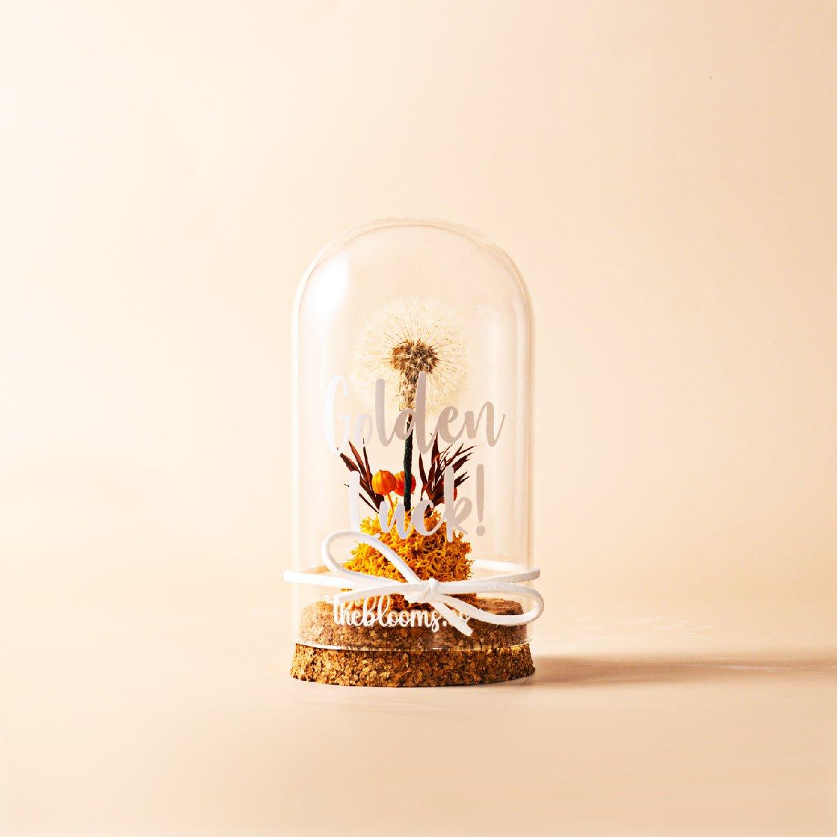 A glass dome containing a white dandelion seed head, orange preserved flowers, and dried leaves on a bed of yellow moss. The dome sits on a cork base with a white ribbon tied around on the glass dome, and the background is a solid light tan color.