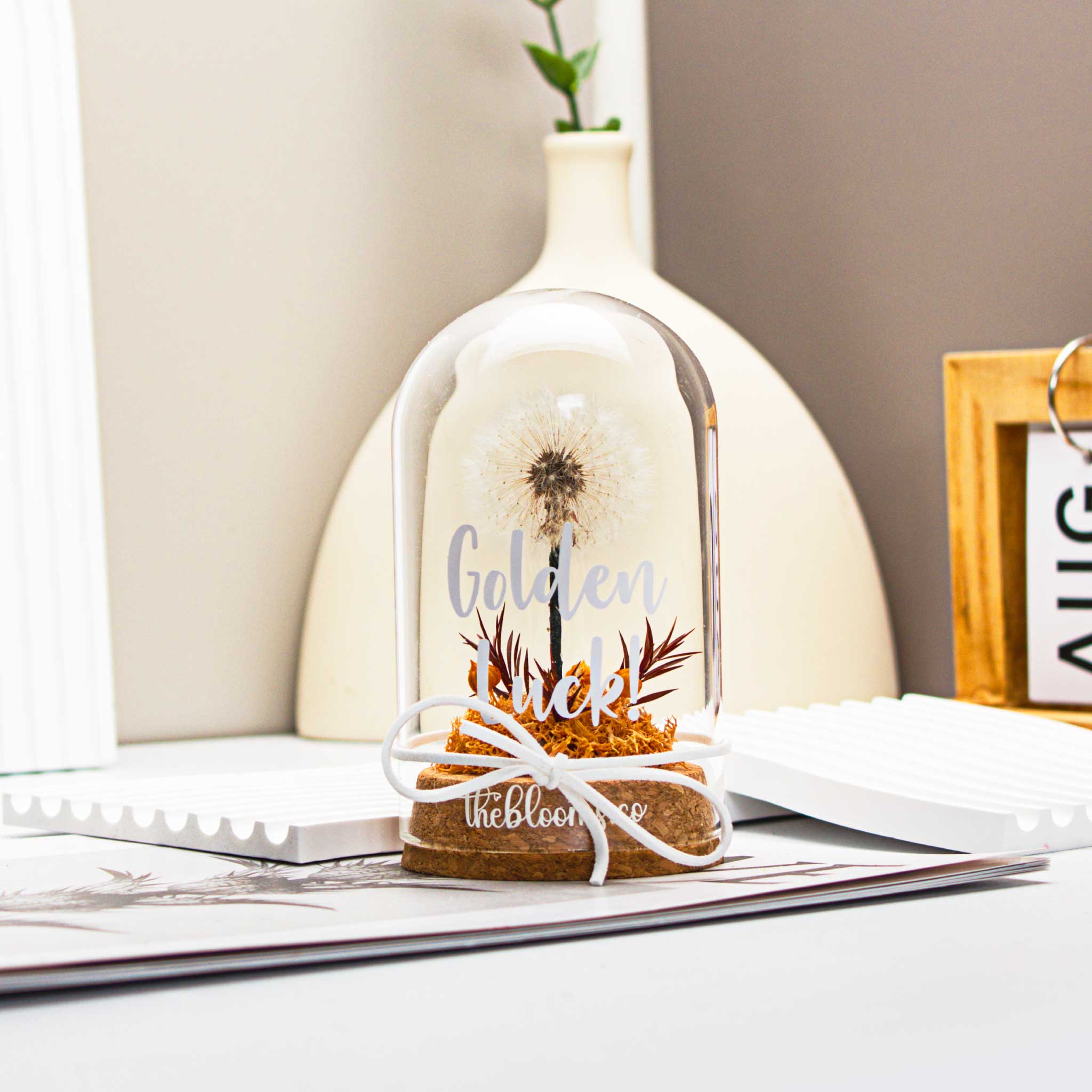 
Glass dome with a single, white dandelion puffball and a small cluster of Orange preserved flowers inside. The dome is on a round wooden base and tied with a white ribbon.