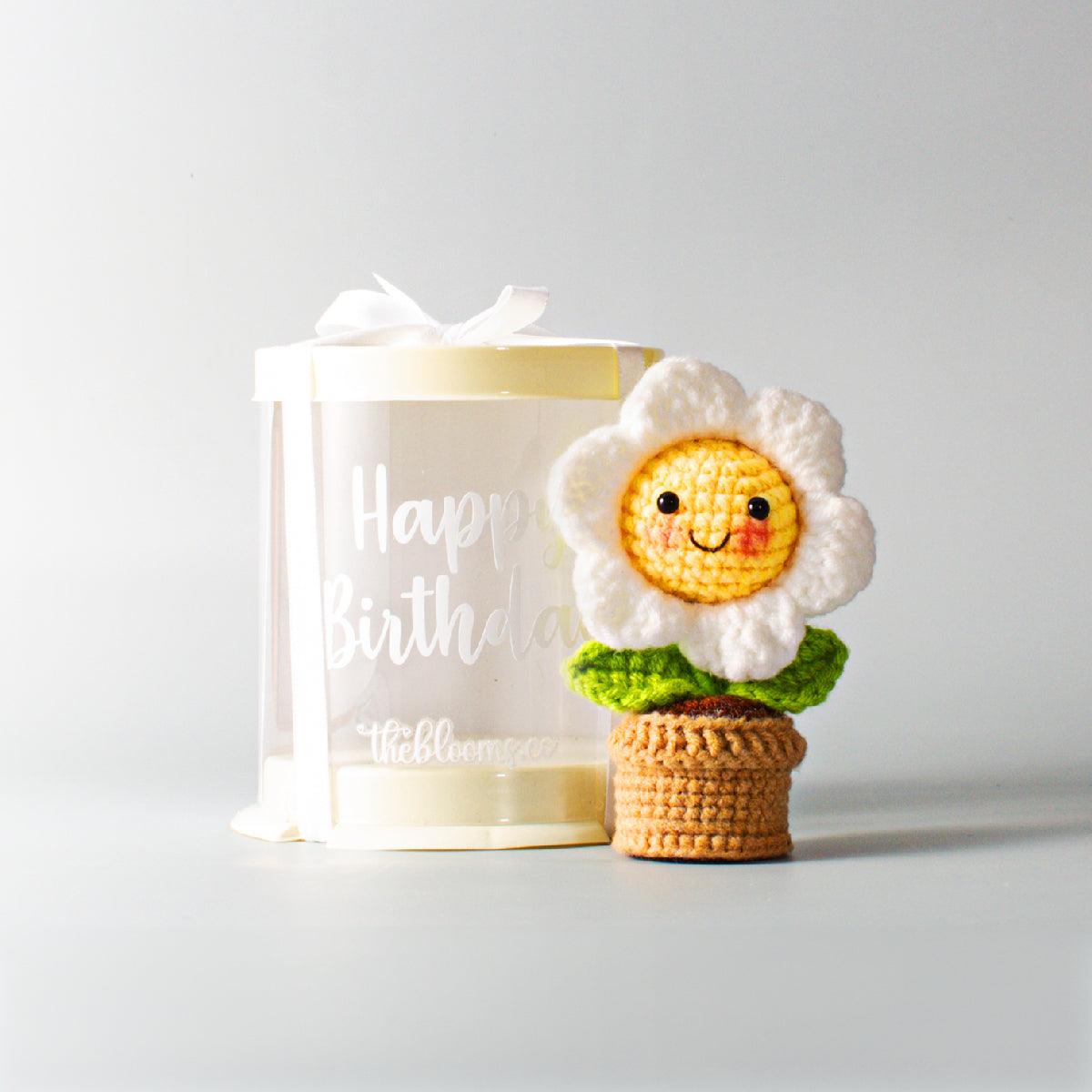 
A small, handmade crochet daisy with a cheerful, smiling face is sitting next to a cylindrical, semi-translucent gift box. The daisy has white petals, a yellow center with rosy cheeks and a happy expression, and green leaves, and is "potted" in a small, brown crochet container. The gift box has a pale yellow top and base, is tied with a white ribbon, and has "Happy Birthday" and "theblooms.co" printed on its side in white font. Both items are placed on a clean, light gray surface against a simple, light gr