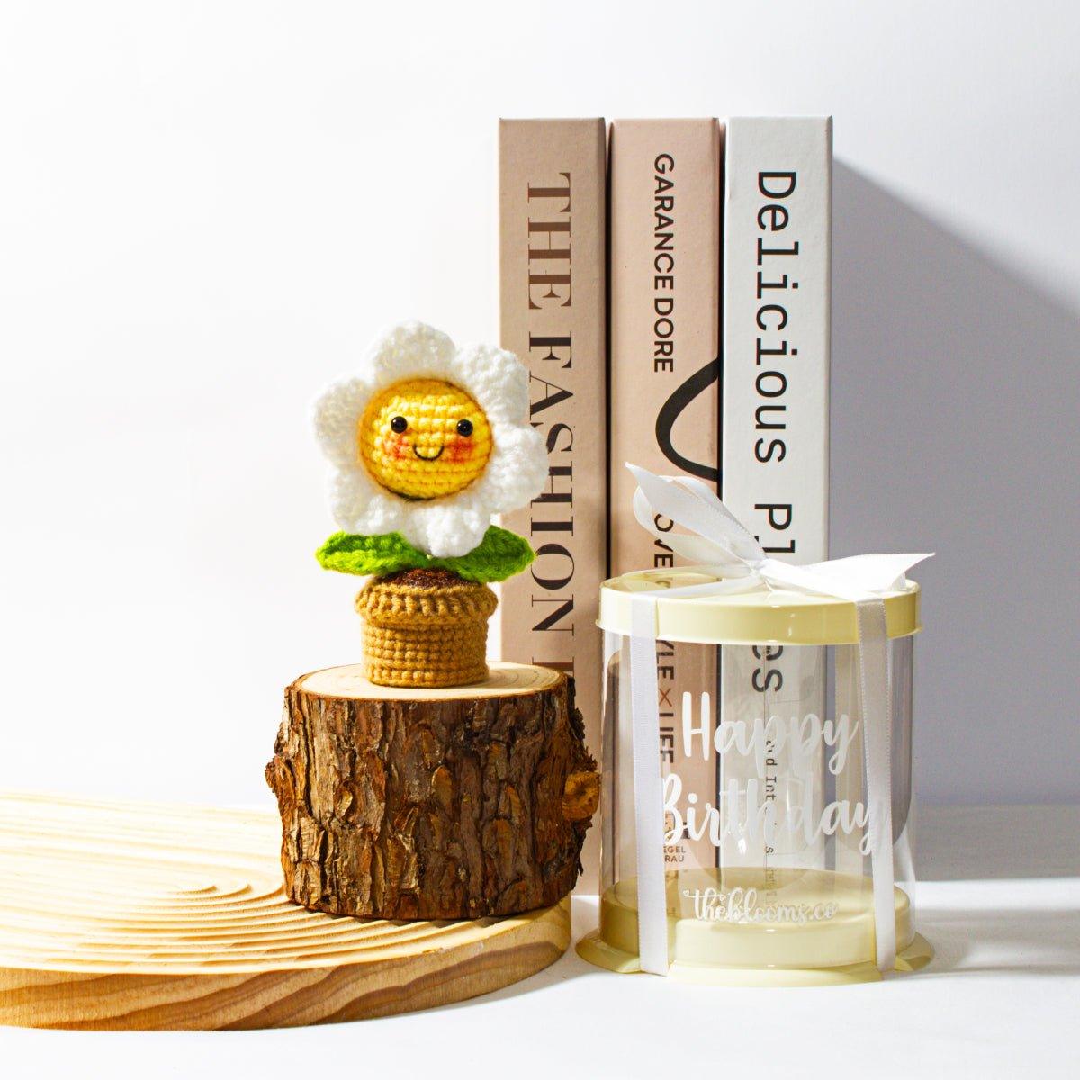 A charming crochet daisy with a smiling face in a small terracotta-colored pot, placed on a thick, natural wooden log slice. Next to it is a cylindrical, semi-translucent yellow gift box with a white ribbon tied on top. The box is decorated with the words "Happy Birthday" in white lettering and the logo "theblooms.co"