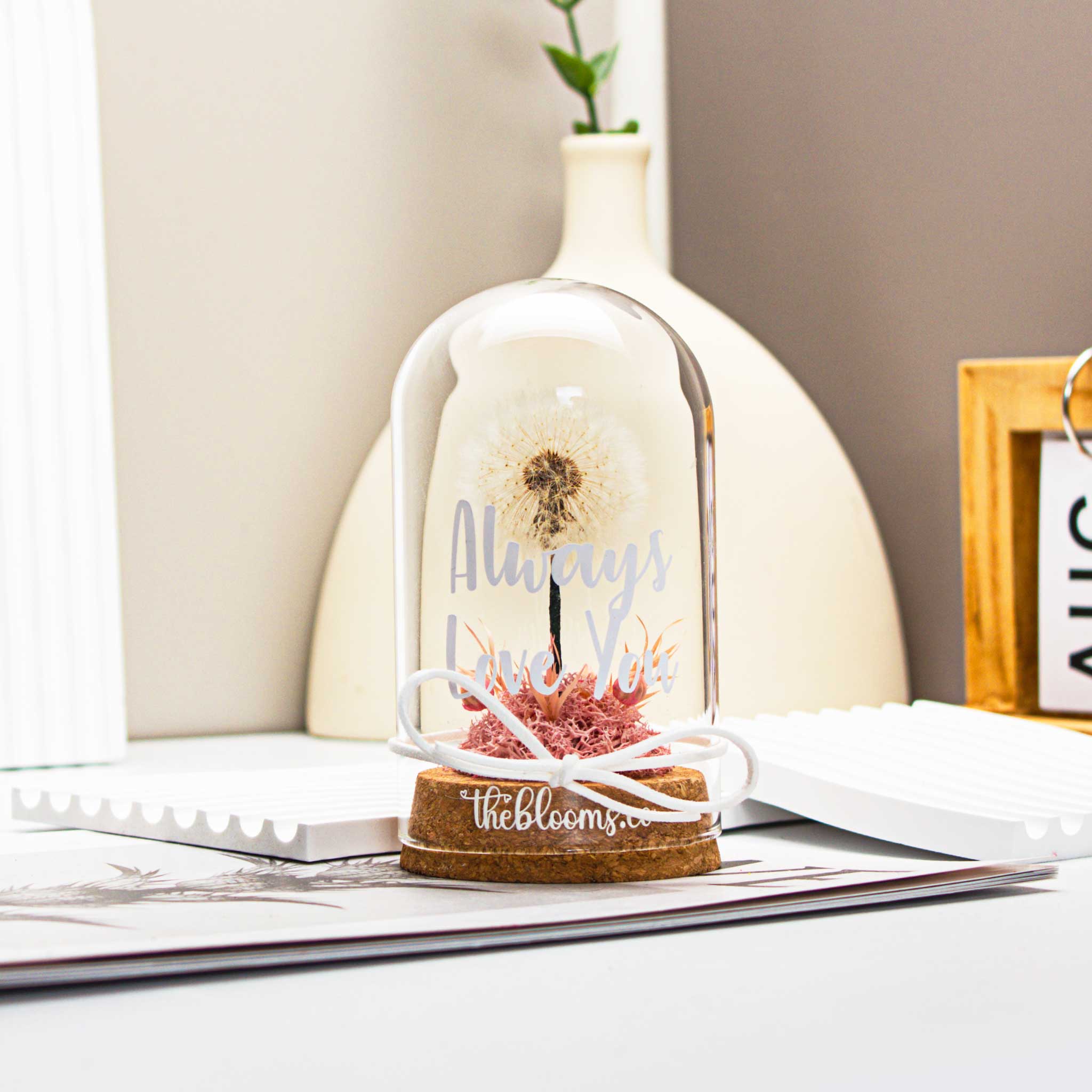 
Glass dome with a single, white dandelion puffball and a small cluster of Pink preserved flowers inside. The dome is on a round wooden base and tied with a white ribbon.